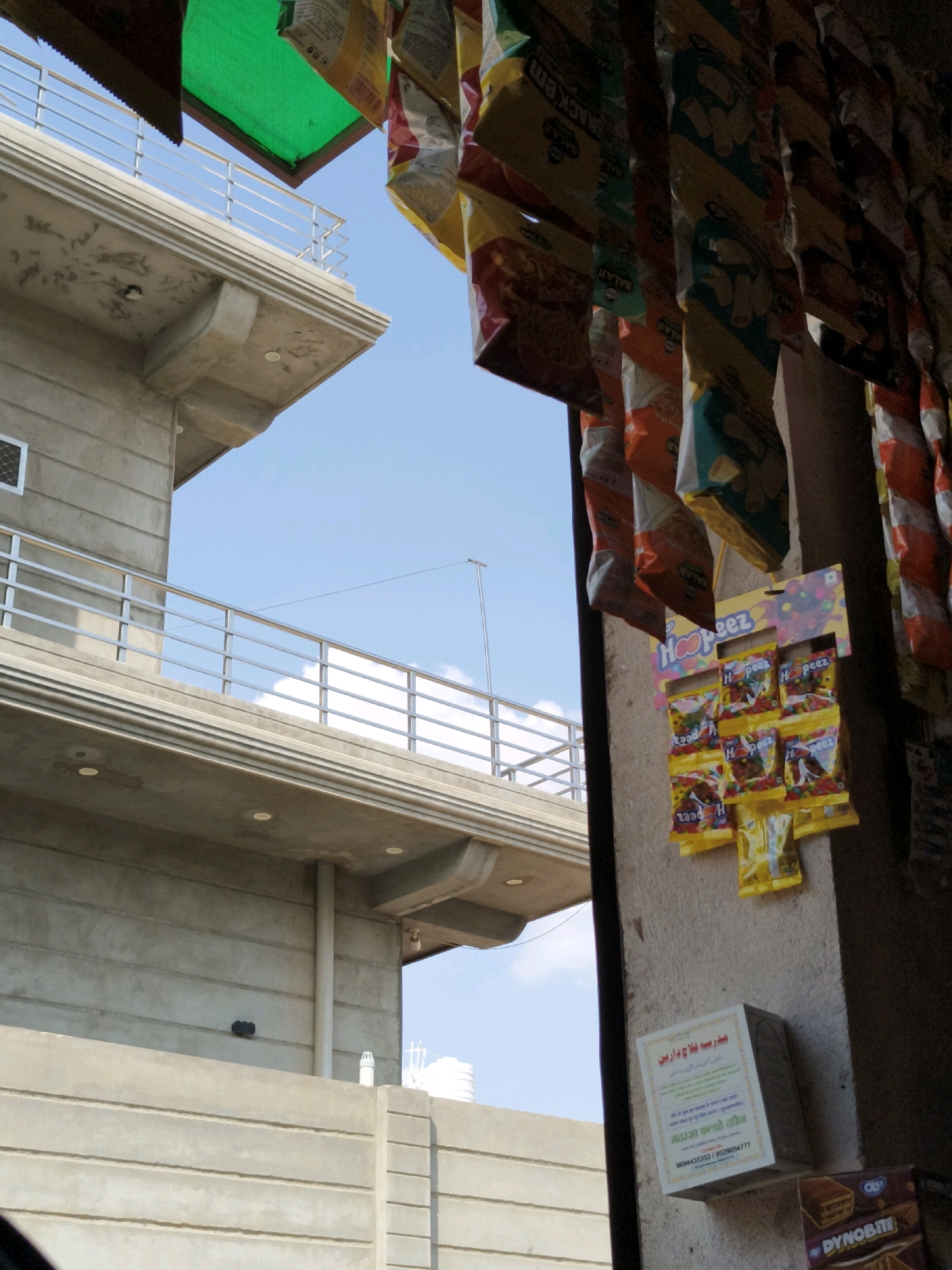 The image shows a street-level view in Taranagar, India. In the foreground, several bags of snacks and candy are hanging from a storefront. To the left, a multi-story building with a concrete facade and a metal railing is visible against a blue sky with some clouds. The building appears to be under construction or recently built. A portion of a white wall with horizontal indentations is in the lower part of the image, and further to the right, a cardboard box with a picture of a chocolate bar is visible. The overall scene depicts a commercial area with elements of both old and new construction.