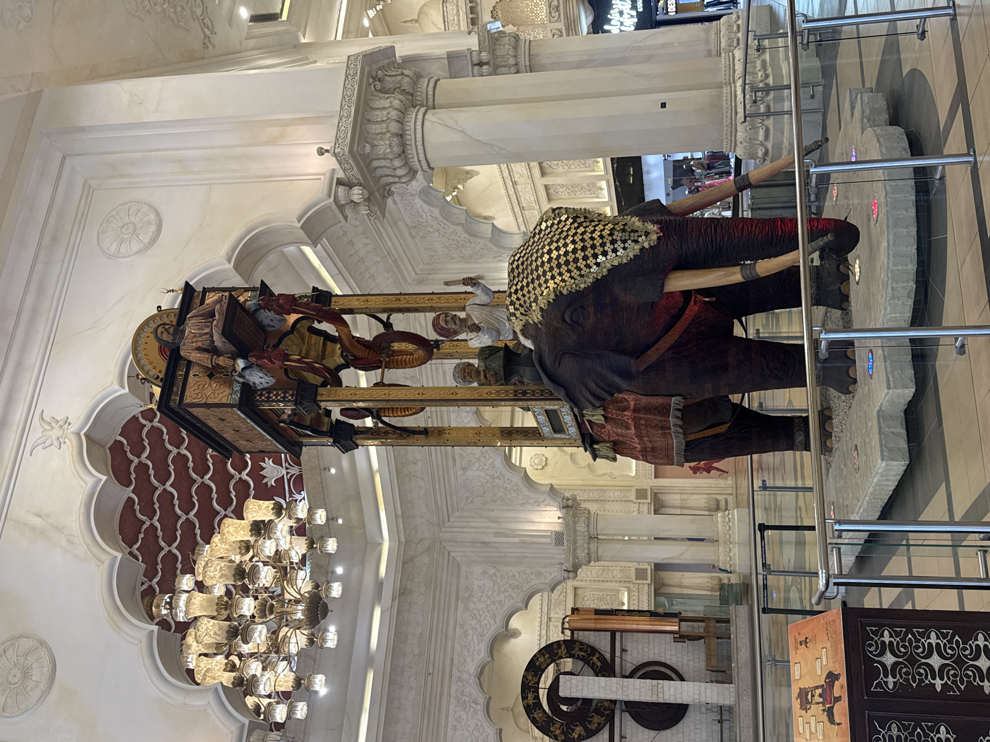 An ornate, life-sized animatronic elephant with a decorative saddle cloth and tusks stands in the center of a large, indoor, white-and-gold public space. The elephant is adorned with a gold and black patterned cloth over its head and a red and gold textile draped over its back. Two figures are seated on the elephant: one in a high structure on its back, and another man in a turban on the elephant's neck. A tall, intricate wooden structure with a clock and further figures rises above the elephant. The background features grand architecture with arched doorways, columns, and elaborate decorative moldings. Above and to the left, a large, multi-tiered chandelier hangs from the ceiling, illuminating the scene. The floor appears to be made of polished tile. The elephant is surrounded by a glass barrier. A sign with an illustration of the elephant and text is visible to the left. The environment suggests a luxurious indoor setting, possibly a mall or hotel lobby, and the architecture's style points to a Middle Eastern or Indian influence. The overall impression is one of grandeur and spectacle, typical of installations found in Dubai, United Arab Emirates, particularly in high-end commercial or entertainment venues.