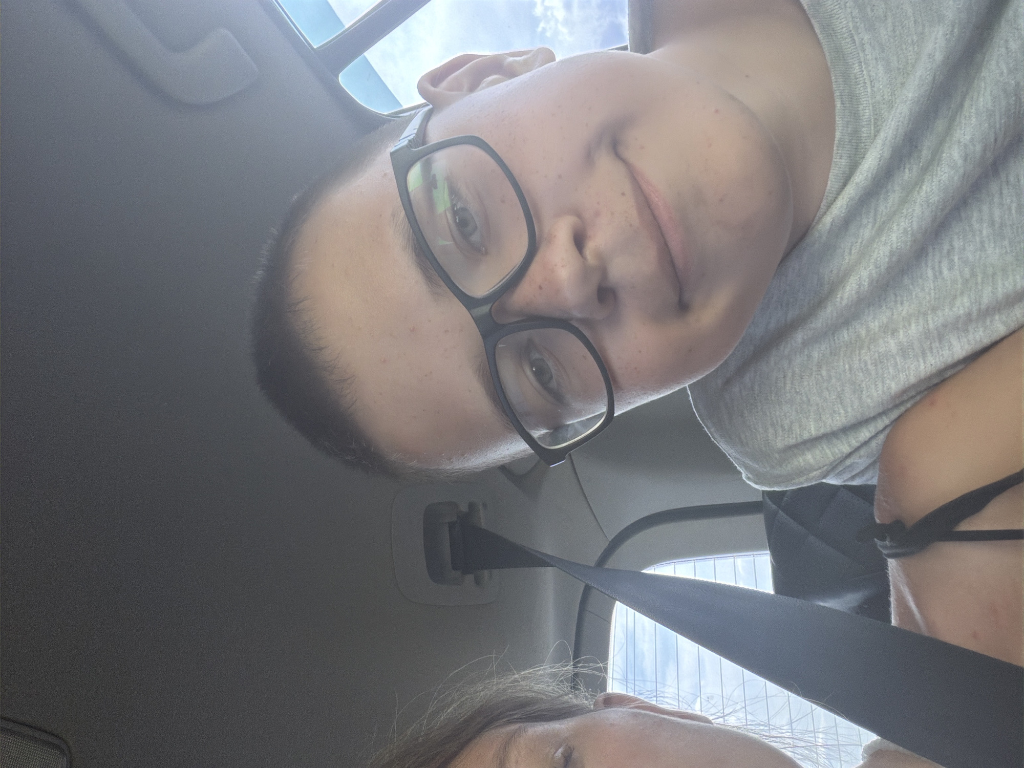 A selfie taken from inside a car shows two people. On the right is a young person wearing glasses and a grey t-shirt, smiling with their mouth slightly open. On the left, only the face of another person is partially visible, looking downwards. The background shows the interior of the car, including a seatbelt and a window with a view of the sky and what appears to be a treeline. The image is taken during the day.