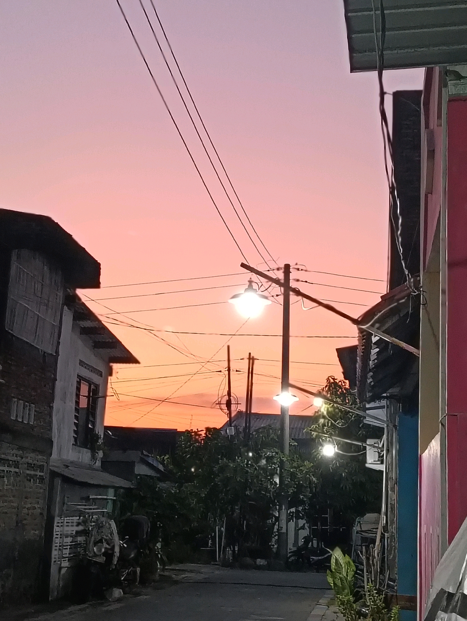 The image depicts a street scene at dusk in Semarang, Indonesia, characterized by a vibrant pink and orange sky. In the foreground, a narrow street is flanked by buildings on either side. To the left, a two-story house with a brick lower level and a white-painted upper level is visible. The upper level has boarded-up windows. Further down the street, foliage and trees are present. On the right, a building with a red facade is partially visible, with a blue trim. Overhead, a network of electrical wires crisscrosses the sky, supported by utility poles. Two streetlights illuminate the scene, one casting a bright glow on the left side of the frame and another with a softer light further down the street. The overall atmosphere is that of a quiet, residential street transitioning from day to night.