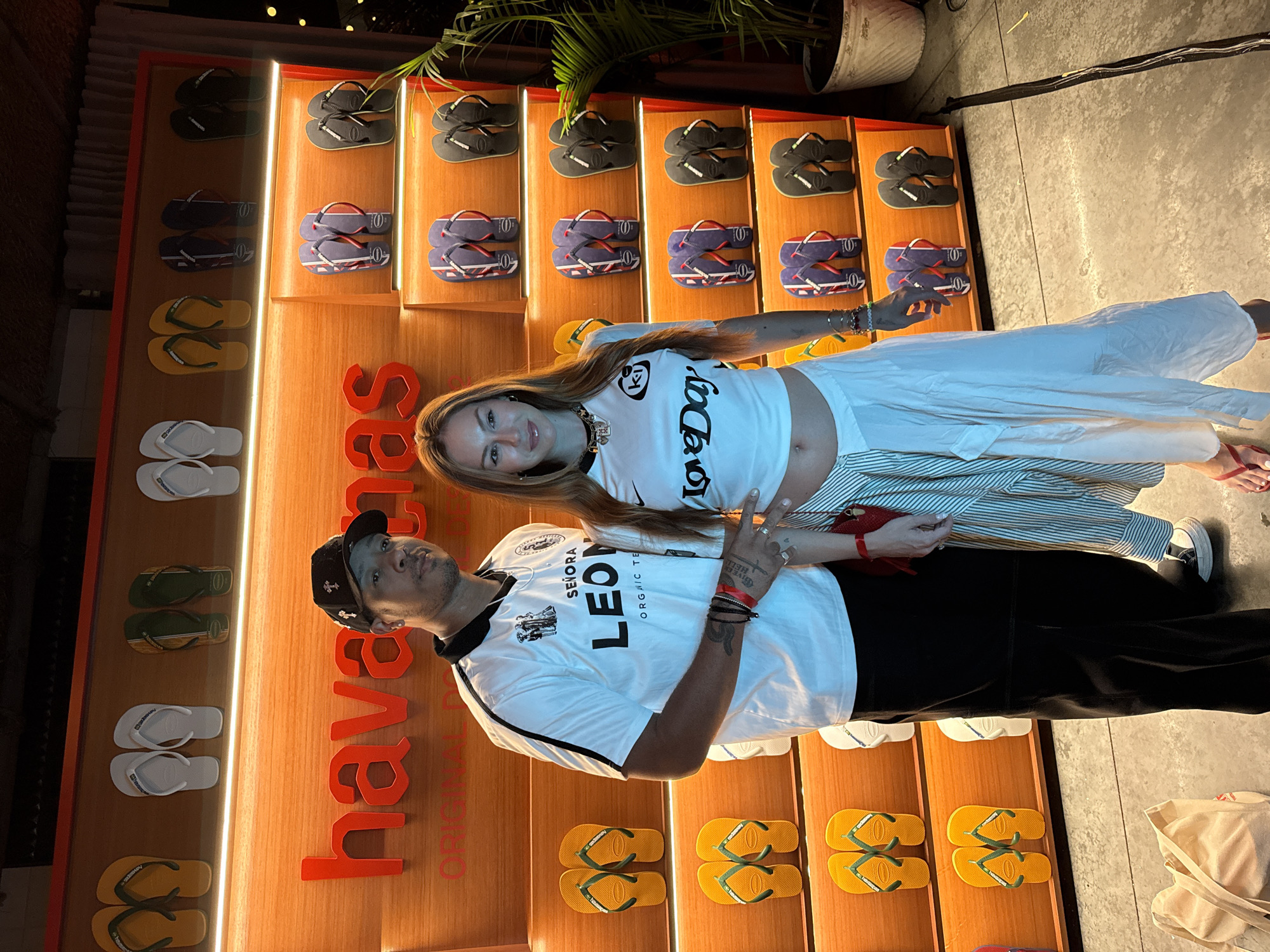In Los Angeles, California, United States of America, a man and a woman stand in front of a display of Havaianas flip-flops. The display wall is made of orange wood and features shelves stacked with various colored Havaianas. The man, wearing a white t-shirt with black trim, a black baseball cap, and black pants, has his right arm around the woman. The woman, wearing a white crop top with "LOVE DAY" printed on it, a long white skirt with striped panels, and red flip-flops, has her left arm around the man and her right hand is making a peace sign. Both are looking towards the camera. The environment appears to be an indoor retail space or event.