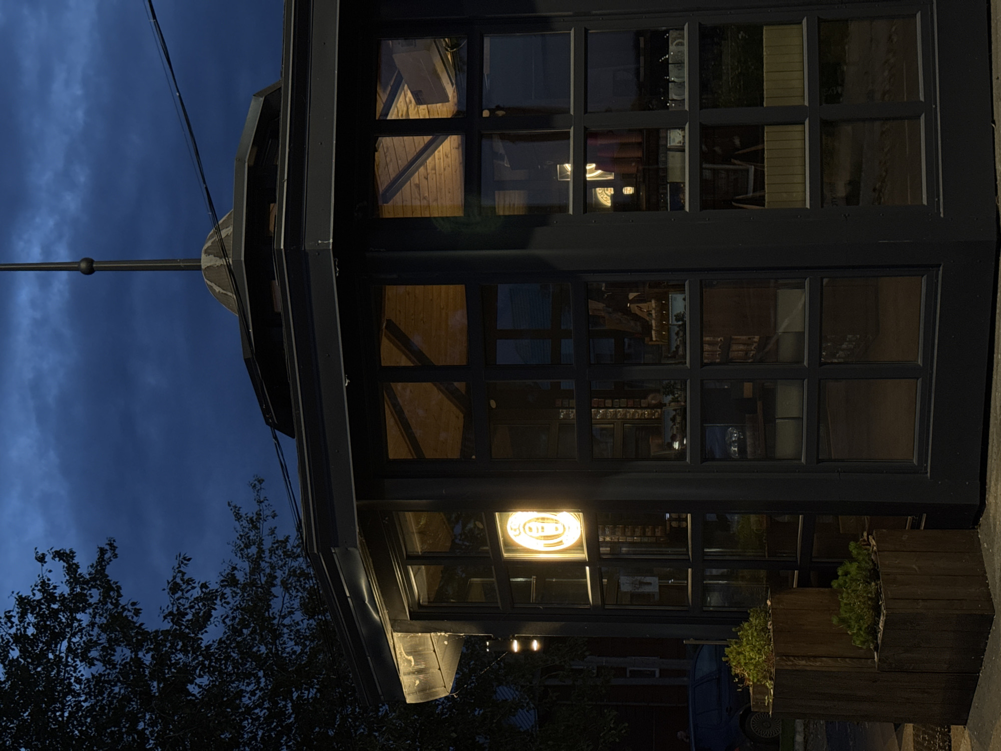 A multi-sided, dark-colored kiosk structure with large glass windows is pictured at dusk. The kiosk has a dark, peaked roof with a thin, dark spire extending from its apex. A bright yellow neon sign in the shape of a cup is visible through one of the windows. Inside the kiosk, the warm glow of lights illuminates wooden interior paneling and what appears to be merchandise or display shelves. Two wooden planter boxes containing low-lying green plants sit in front of the kiosk. To the left of the kiosk, a dark-colored car is parked on the street. The background consists of a dark, cloudy sky and the silhouette of a tree. The scene is located in Votkinsk, Russia.