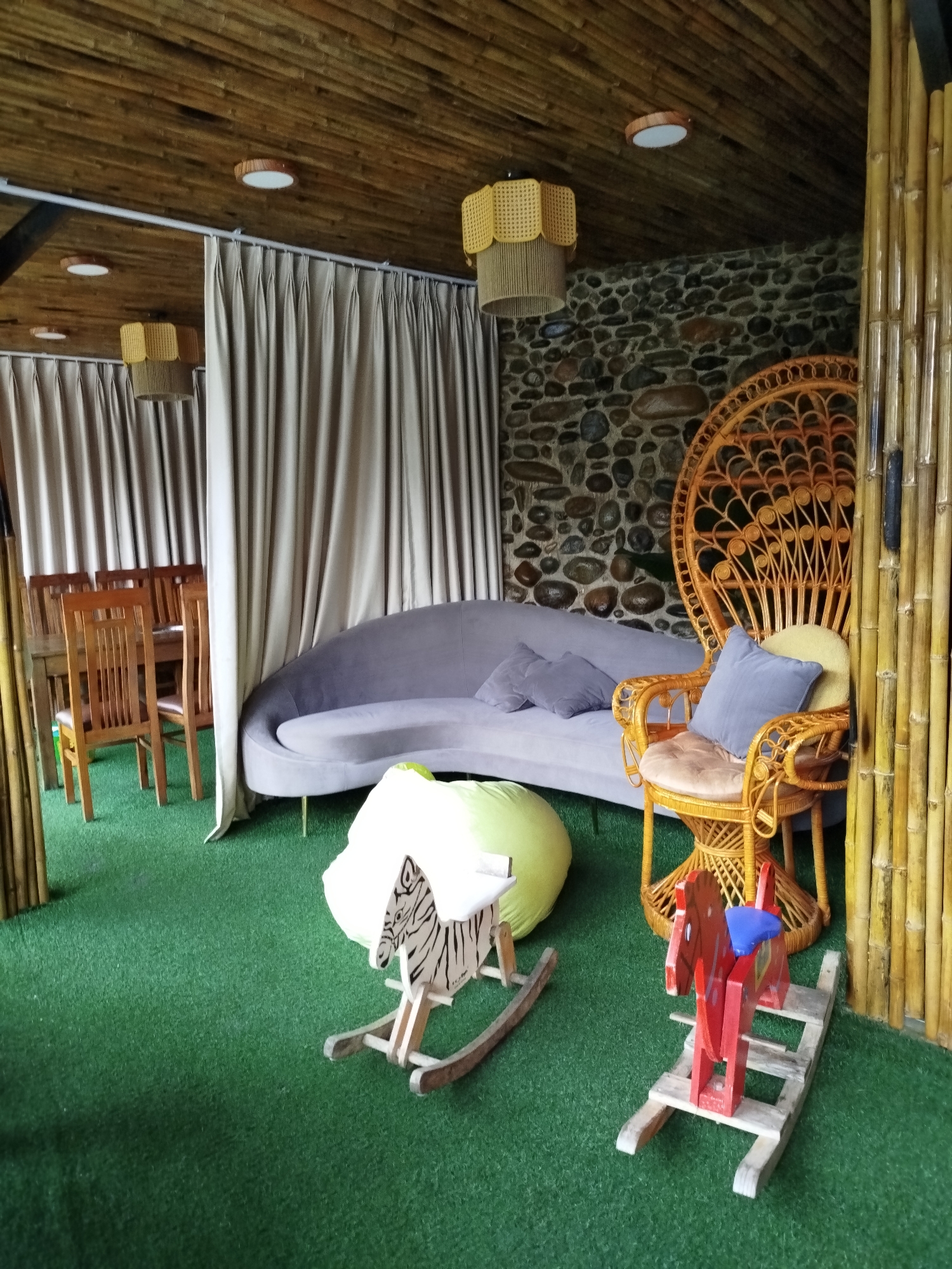 The image depicts a room with a bamboo ceiling and artificial grass flooring. The room is in Da Lat, Vietnam. A grey curved sofa with two purple pillows sits against a stone wall, next to a rattan peacock chair with a yellow cushion and a blue pillow. In the foreground are two rocking horses, one white with black stripes and the other red with a blue seat. Curtains partially enclose a dining area with a wooden table and chairs. A yellow lampshade hangs from the ceiling. Bamboo columns frame the right side of the room.