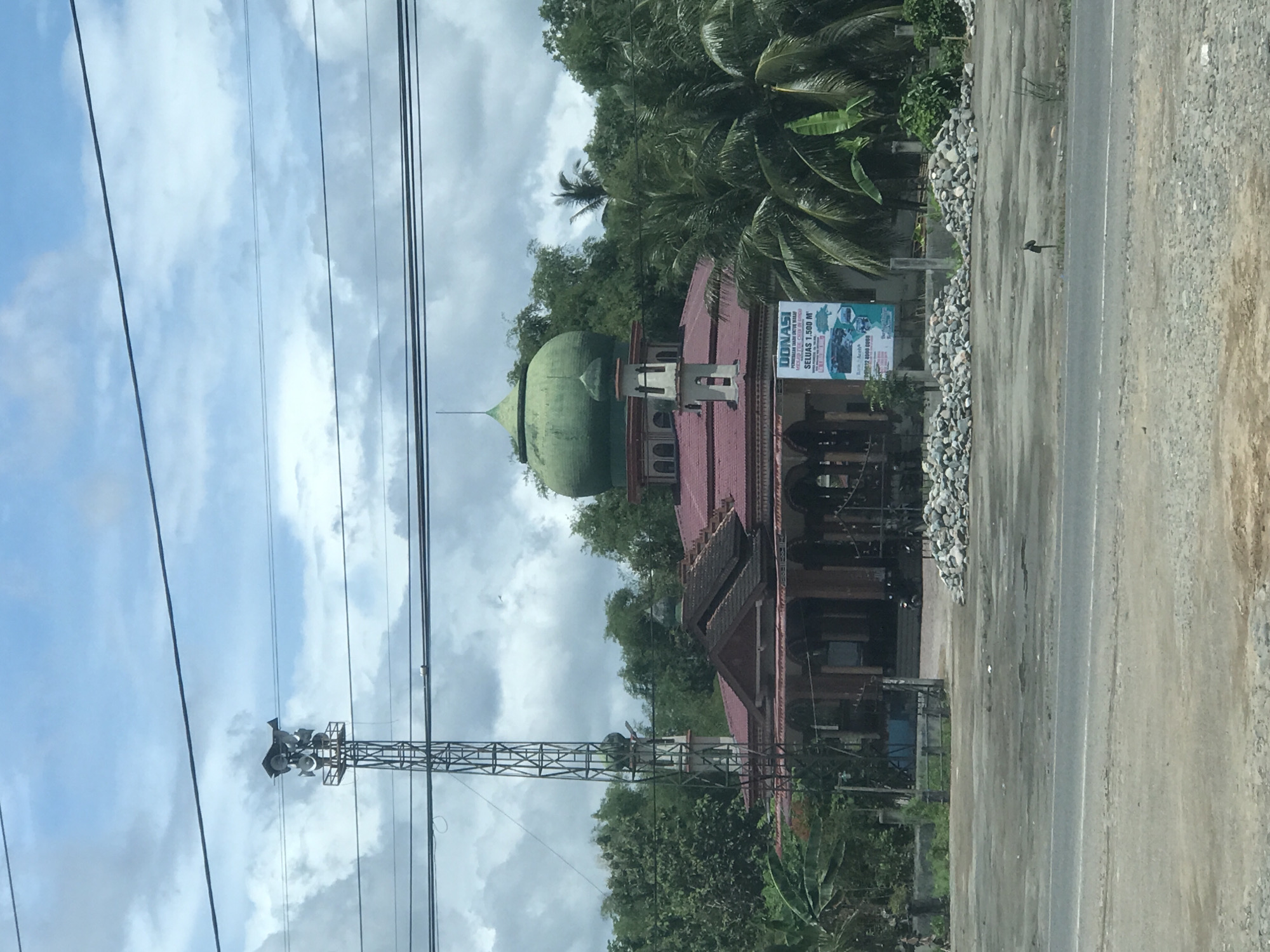 The image shows a mosque in Bulo Gogo, Indonesia, with a green dome and red roof. The mosque is situated next to a road. A communication tower is visible to the left of the mosque. Overhead power lines stretch across the sky, which is cloudy with patches of blue. A billboard is attached to the side of the mosque. Trees and other vegetation are present in the background. The ground is a mixture of pavement and gravel.