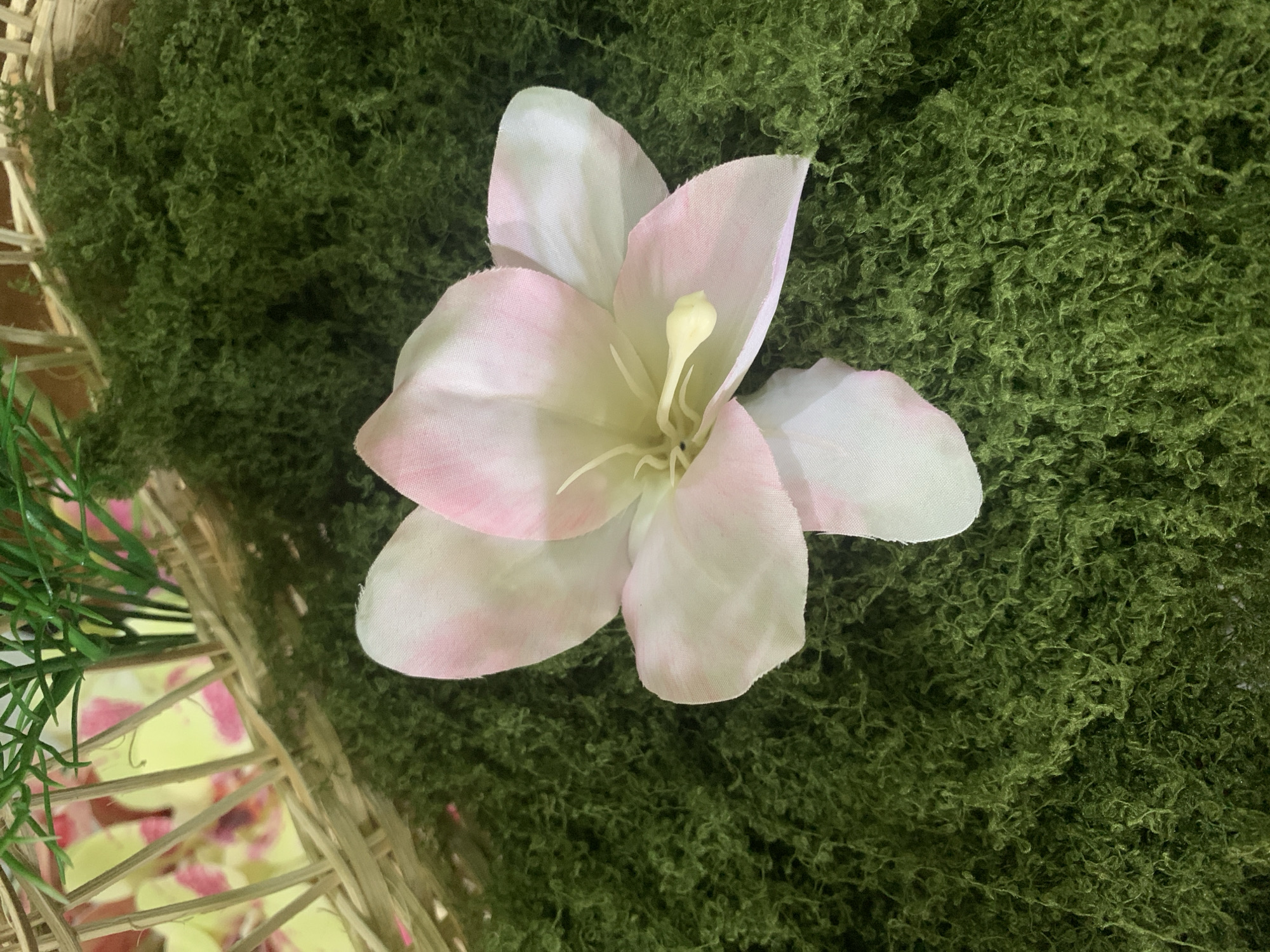 A close-up shot focuses on a light pink and white artificial flower resting on a bed of vibrant green moss. The flower, with its delicate petals and yellow stamen, is centrally positioned in the frame. To the upper left, a portion of a woven basket is visible, containing what appears to be a yellow and pink floral arrangement and some green artificial grass. The setting is an indoor environment, possibly a craft or decorative display, with no specific identifiable landmarks of Kuala Pembuang, Indonesia.