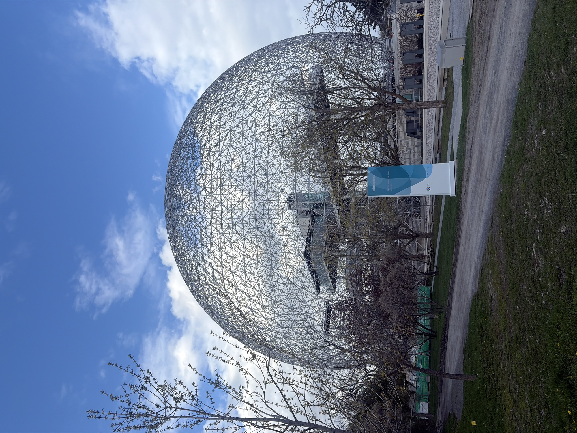 The image presents the Montreal Biosphere under a blue sky with scattered clouds. This geodesic dome structure, located in Montreal, Canada, is made of a network of interconnected metal rods forming numerous triangular shapes. Trees with bare branches partially obscure the Biosphere. Adjacent to the Biosphere, there is a pathway with grass and a signpost. Buildings are visible behind the Biosphere. The overall perspective is from a low angle, looking upwards toward the Biosphere against the sky.