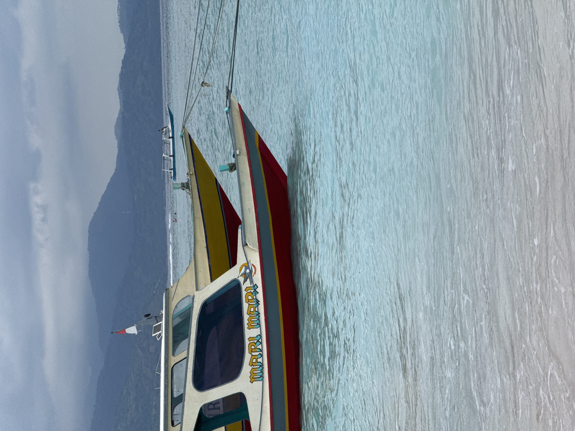 A color photograph shows a boat, with the name "MARI MARI" emblazoned on its side, floating in water. The boat has a red and yellow trim. In the background, a hazy mountain range is visible. The location is in the city of Unknown, Indonesia. The sea surface appears calm, and the boat seems to be stationary. The sky is overcast. A flag is visible on the boat's mast. The photograph is taken from an angle looking towards the boat and the mountain range.