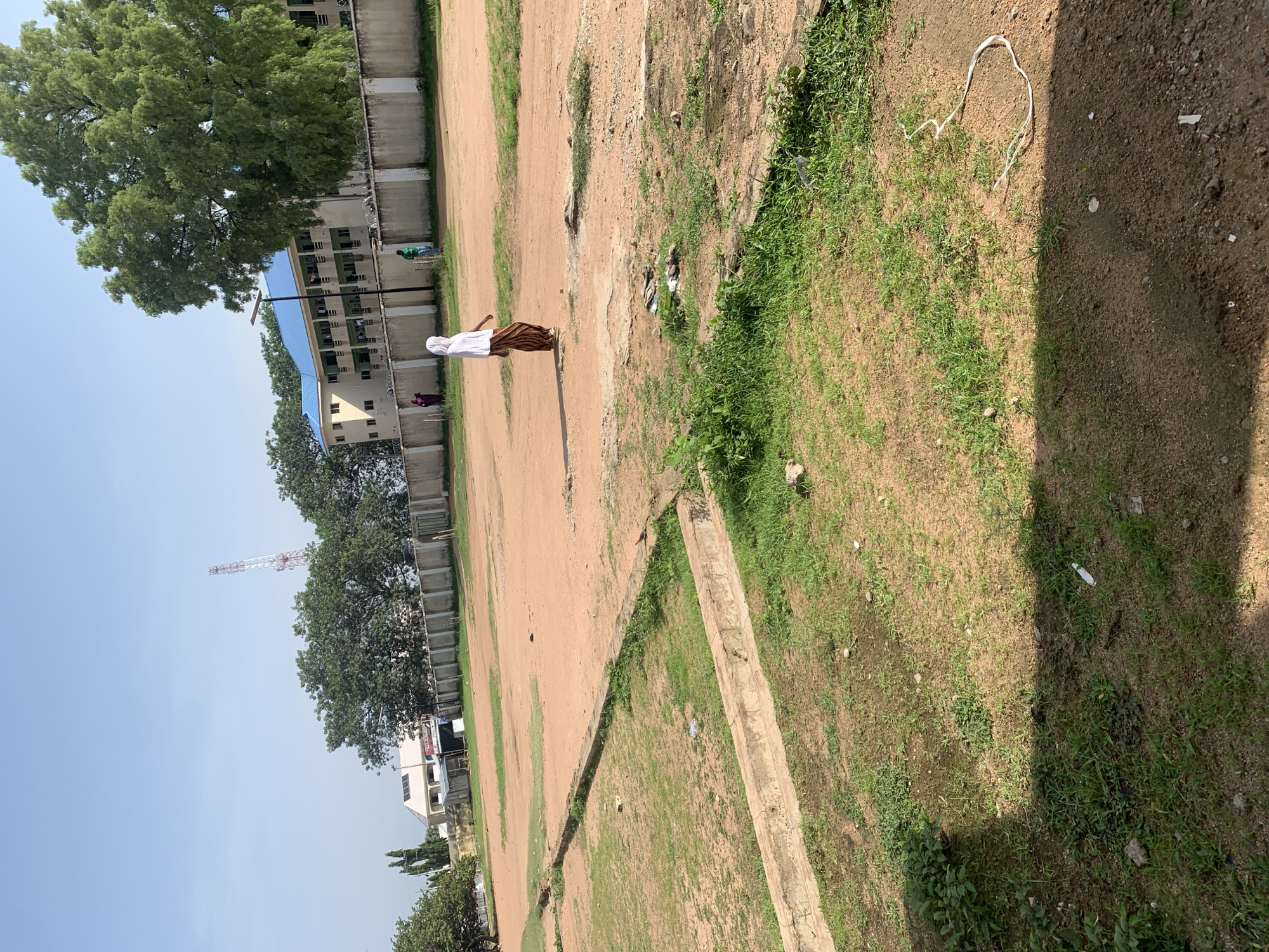 The image depicts an outdoor scene on a clear day, likely in Jalingo, Nigeria. In the mid-ground, a person wearing a white headscarf and a striped skirt walks across a dusty, open field. The field is bordered by a concrete wall topped with a fence. Beyond the wall are buildings with blue roofs and trees, and further in the background, a tall communications tower is visible. The foreground shows patches of dry grass and dirt, with some concrete structures suggesting drainage or borders. The overall impression is of an open area adjacent to institutional buildings, possibly a school or administrative complex.
