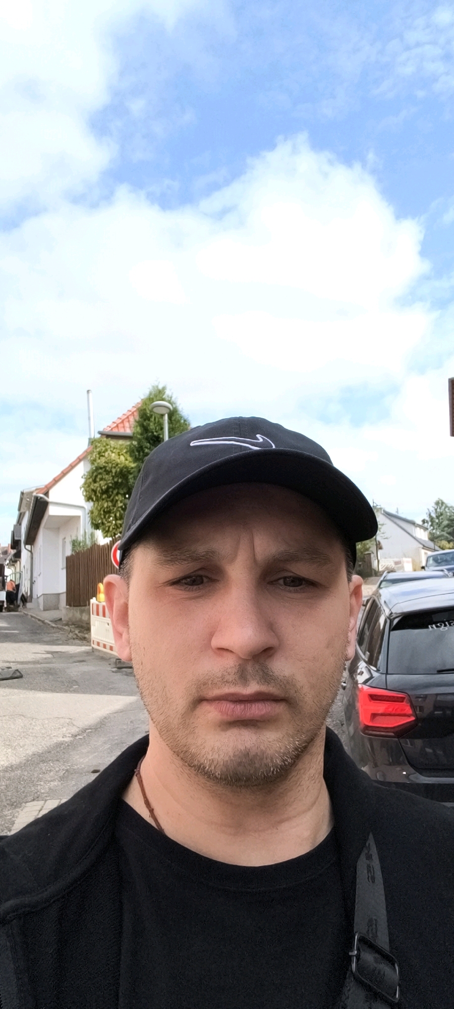 In a street view selfie, a man wearing a black baseball cap and a black jacket is looking directly at the camera. The background shows a street with houses and parked cars. The sky is blue with scattered clouds. On the left side of the image, a white house with a red-tiled roof and a fence can be seen, with some construction barriers in front of it. A dark-colored Audi car is parked on the right side of the street. The scene appears to be in a residential area.