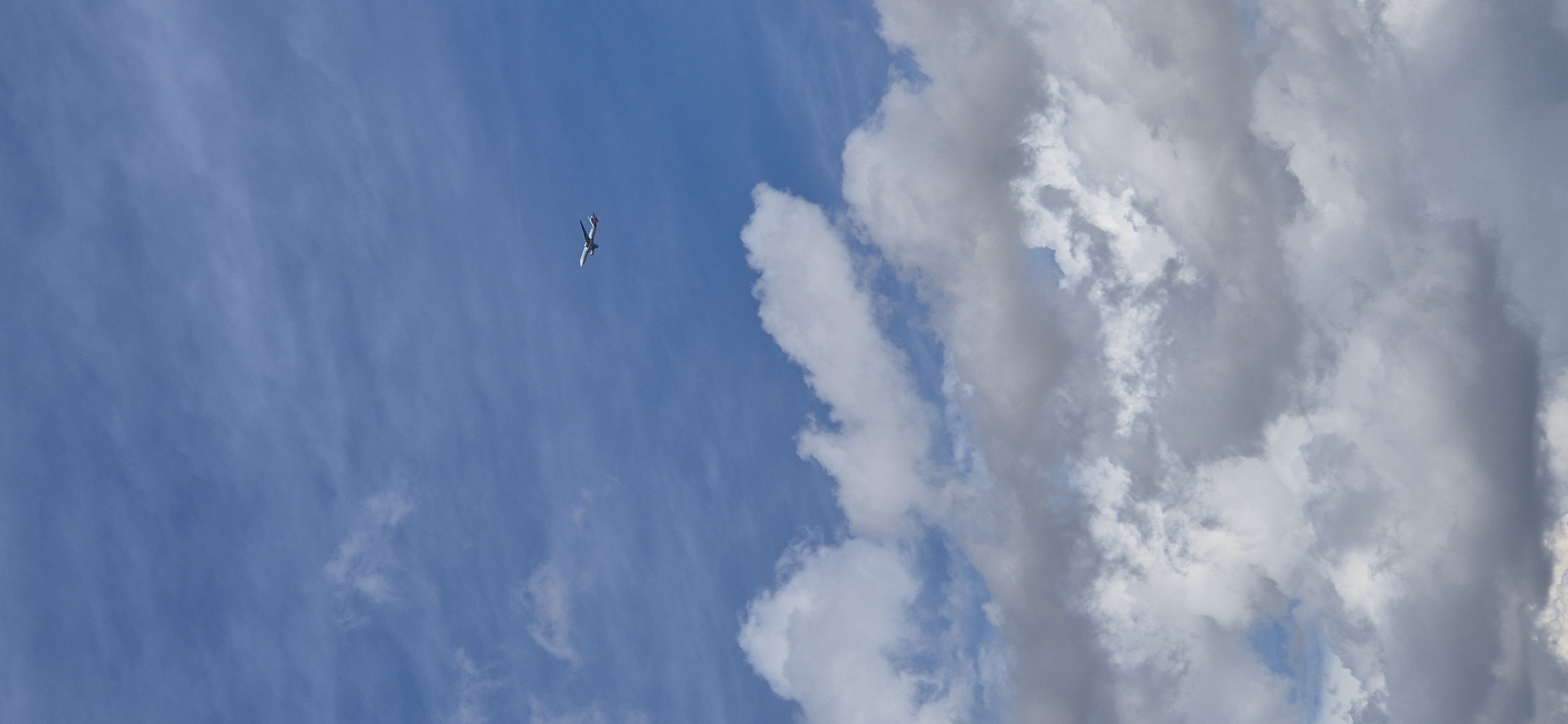 A small commercial airplane is visible in the upper right quadrant of the frame, flying against a backdrop of a blue sky with scattered white and gray clouds. The plane is a white commercial jet with a red stripe along its fuselage and tail. The clouds are varied in density, with some appearing wispy and others more substantial and textured. The lower portion of the image is dominated by denser, darker clouds, suggesting a change in weather. The scene appears to be an aerial view, with no ground-level elements visible. Without further information or context related to Mainz, Germany, it is not possible to determine the specific location within the city.