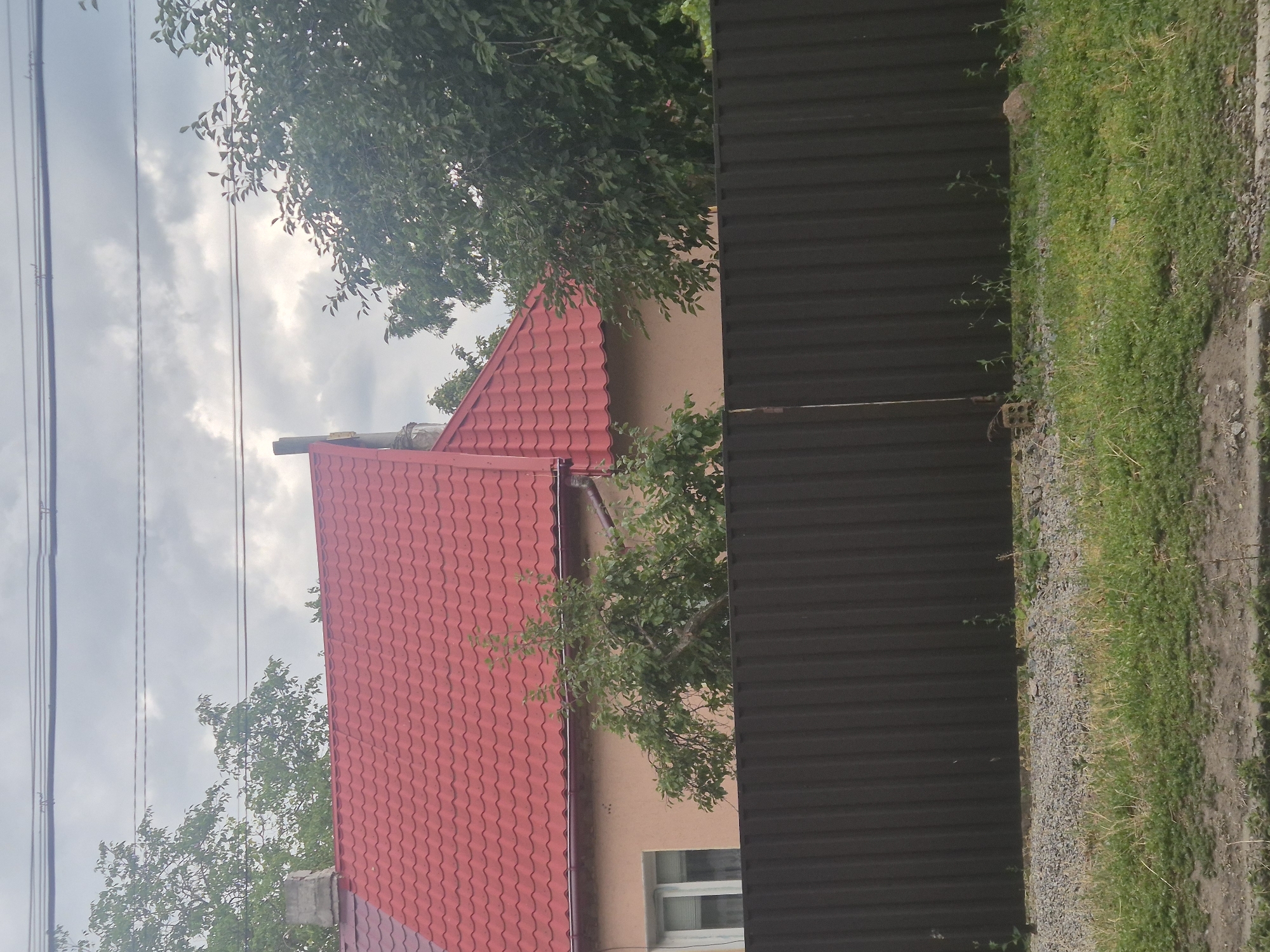 The image shows a residential property with a red corrugated metal roof, a light-colored house, and a dark brown metal fence. The fence runs horizontally across the lower portion of the image. Behind the fence, there are patches of green grass and gravel. Trees are visible on the left and right sides of the property, with the house situated between them. The sky is overcast with gray clouds. The scene appears to be a typical residential area in Sozonivka, Ukraine.