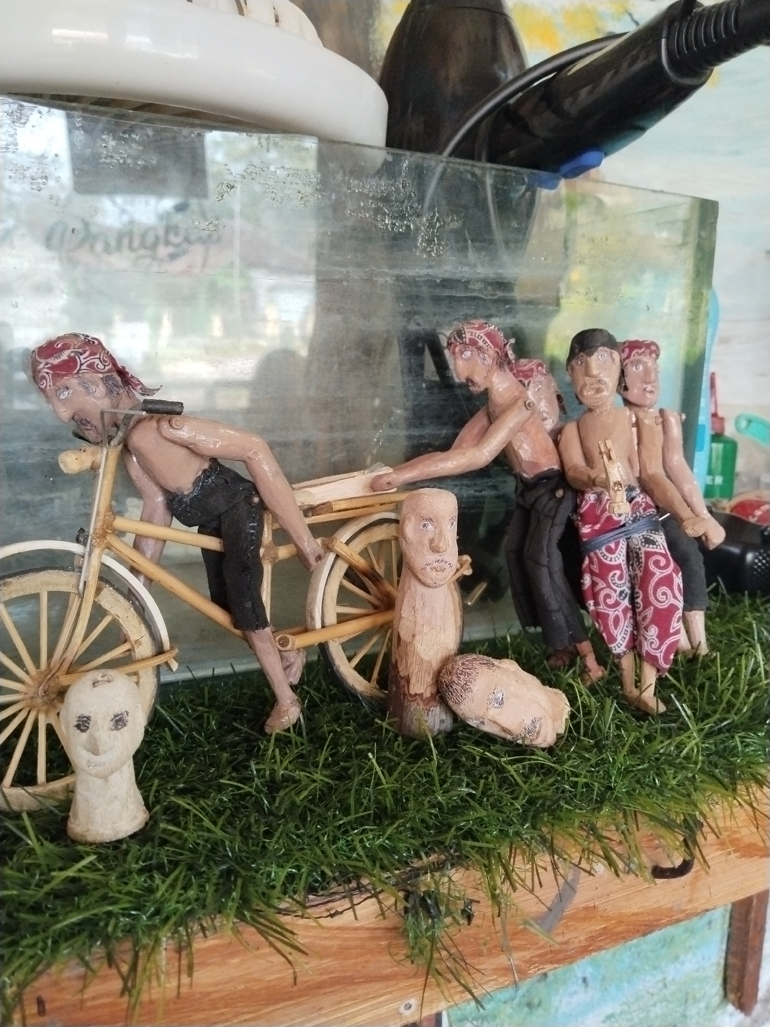 This image presents a close-up view of a miniature diorama, likely a cultural craft display from Jetis, Indonesia. The central focus is a group of four handcrafted figurines, appearing to be made of wood or clay, depicted in the act of pushing a two-wheeled wooden cart or bicycle.
One figure is positioned at the front of the cart, leaning forward and holding its structure, with a red patterned bandana on his head and dark trousers. Immediately behind him, another figure also pushes the cart's frame, wearing similar attire. Two more figures stand to the right, behind the cart. One of these wears distinctive red and white patterned shorts or a sarong and holds a small, carved stick-like object in his hand, while the fourth figure is partially obscured. All visible figures have head coverings. Their postures convey effort or struggle.
The miniature cart itself is intricately detailed, with wooden wheels and spokes. The figurines are placed on a vibrant green artificial grass mat, which rests on a natural wood plank surface. In front of the cart, on the grass, are two detached carved wooden heads or busts; one is upright, and the other lies on its side.
Behind the entire display is a clear, somewhat dirty or aged glass enclosure, possibly a display case or an old aquarium. Through this glass, blurred background objects are visible, including a prominent white plastic object on the left and a black appliance, possibly a hairdryer, on the right. Faint, blurry text, appearing to start with "Dongkrak," can be discerned through the glass on the left side, though it's largely illegible. The scene depicts a narrative of manual labor or a journey.