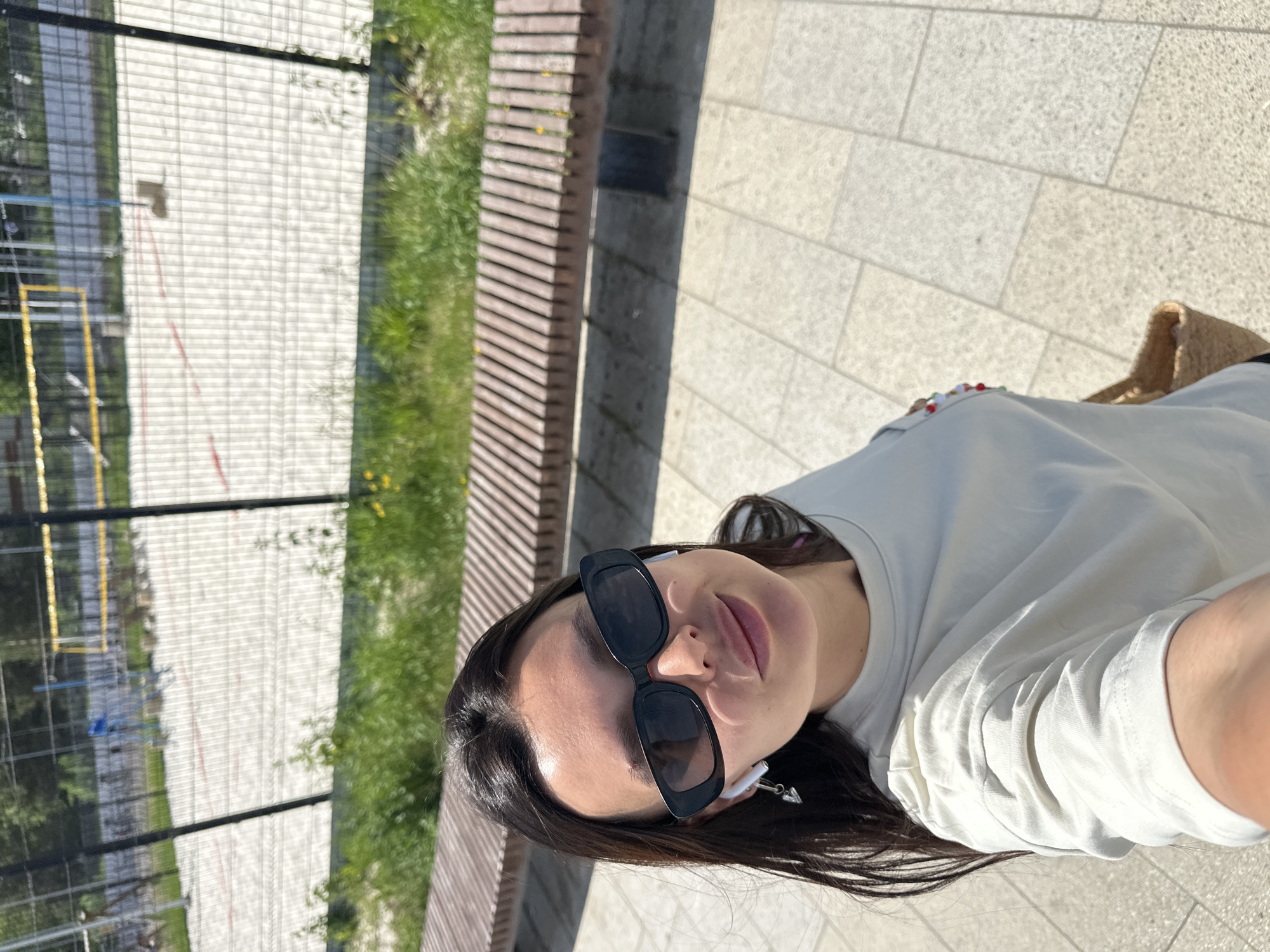 A woman wearing sunglasses and a light-colored shirt is taking a selfie outdoors in Chaikovsky, Russia. She is sitting on a bench next to a sports field with a metal fence. The ground is paved. A straw bag is visible beside her.