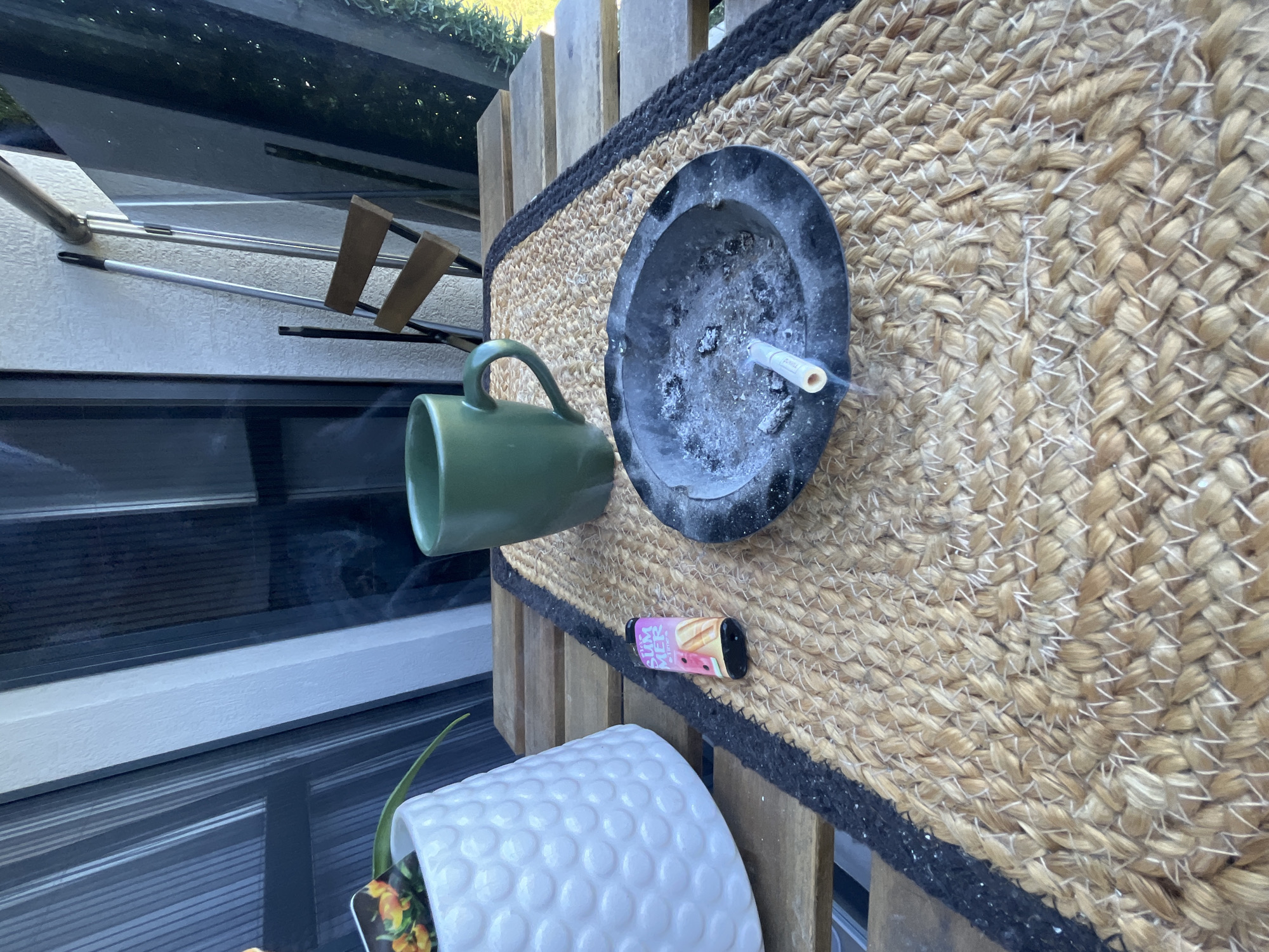 The image depicts a cluttered outdoor scene, possibly a balcony or patio. A black ashtray containing a cigarette butt sits on a woven mat with a black border, along with a disposable vape device. Next to the ashtray is a green mug and a white plant pot. In the background, there's a window or glass pane, reflecting light and suggesting an outdoor setting. Given the request to include the location, and without explicit information within the image, I can only assume this scene is happening in Varna, Bulgaria. The scene suggests a casual setting, potentially someone taking a break.
