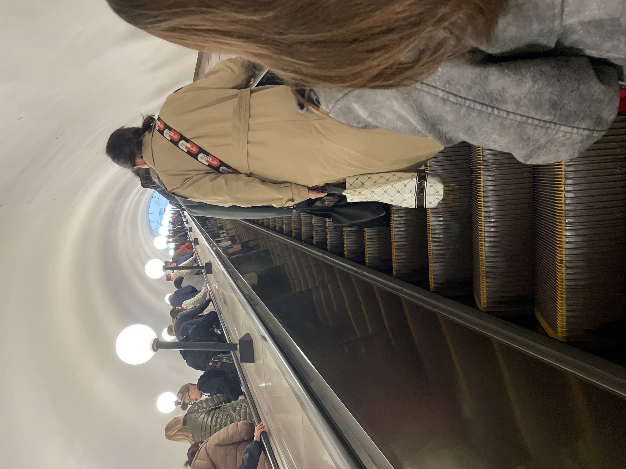The image shows an escalator in the Moscow Metro, Russia, with several people riding it. The escalator is inside a long, curved tunnel with white walls and evenly spaced spherical light fixtures. Passengers are wearing various jackets and coats. A woman is near the top of the escalator. She is wearing a trench coat, black bag, and a purse with a white and black pattern. Other passengers are visible further down the escalator.