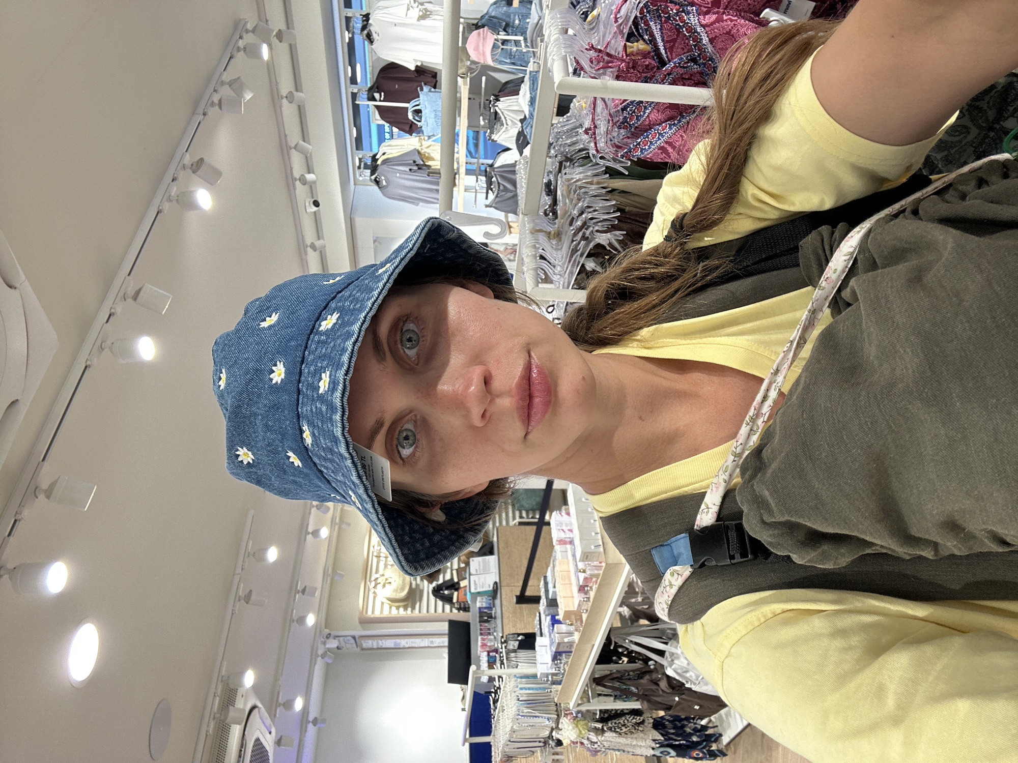 A woman wearing a denim bucket hat with floral embroidery, a yellow t-shirt, and a baby carrier stands indoors in what appears to be a retail store in Fethiye, Turkey. The store has clothing racks filled with various items behind the woman, as well as shelving units with merchandise. Overhead lighting is present.