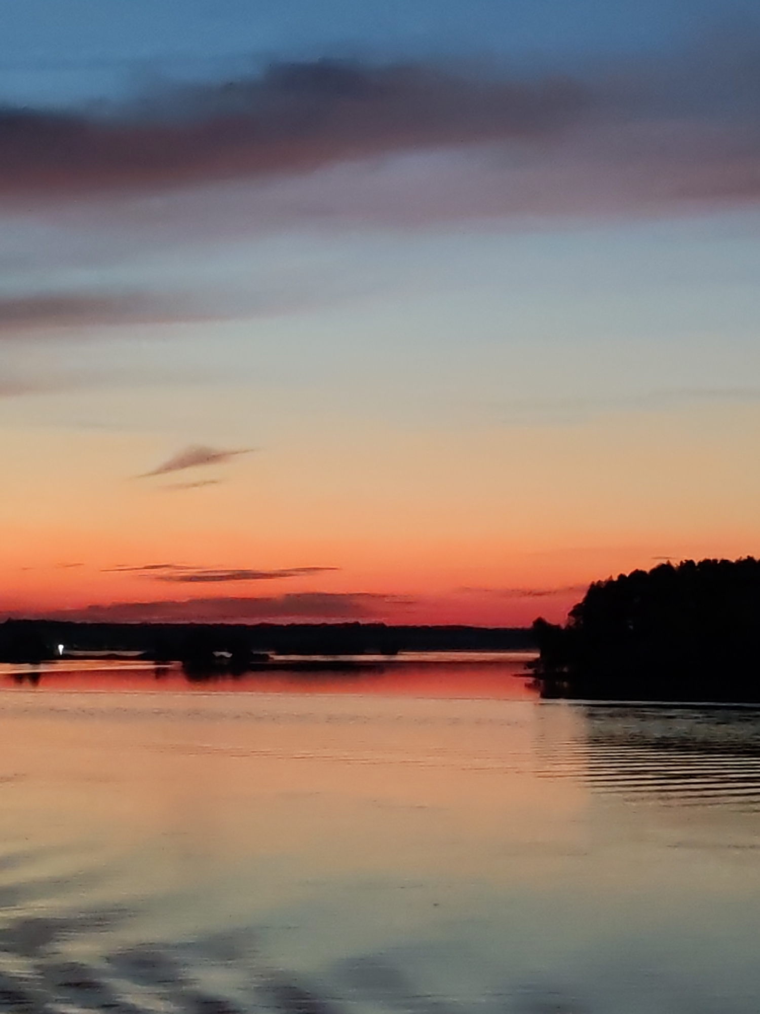 A sunset over a body of water features a sky with bands of blue, pink, and orange. The horizon line is marked by a dark silhouetted tree line, with a sliver of orange light glowing above it. The water reflects the colors of the sky and the silhouetted land. Ripples on the water's surface create a textured effect. The scene is devoid of any people or man-made structures, except for a faint light on the distant horizon, suggesting a possible building or vessel.