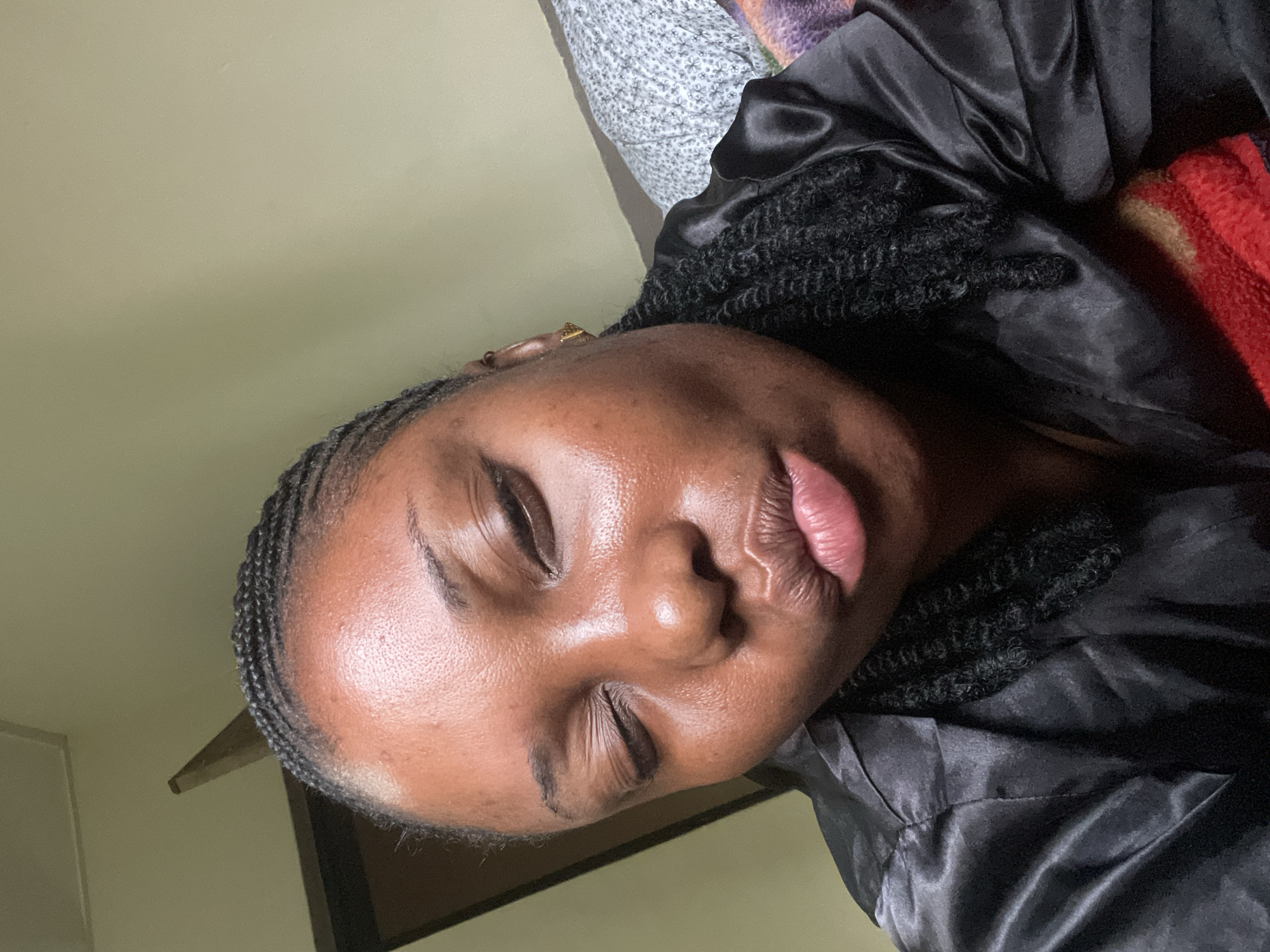 A close-up portrait, oriented sideways, features a person with dark skin resting indoors in Jos, Nigeria. The individual's eyes are closed, suggesting sleep or deep rest, and their lips are slightly parted, revealing a soft pink hue. Their skin appears smooth and has a natural sheen, particularly on the forehead and cheeks. Braided hair, styled as cornrows along the hairline and thicker twists further back, frames their face. A small golden earring is visible on one earlobe.
The person is wearing a black, shiny garment, likely made of satin or a similar material, which covers their shoulders and chest. In the background, a plain, light olive-green wall is visible, along with glimpses of a white patterned fabric (possibly a pillow or sheet) and a folded red or orange textured fabric, likely a blanket or towel. The overall impression is one of peaceful serenity within a comfortable indoor setting. There is no visible text in the image.