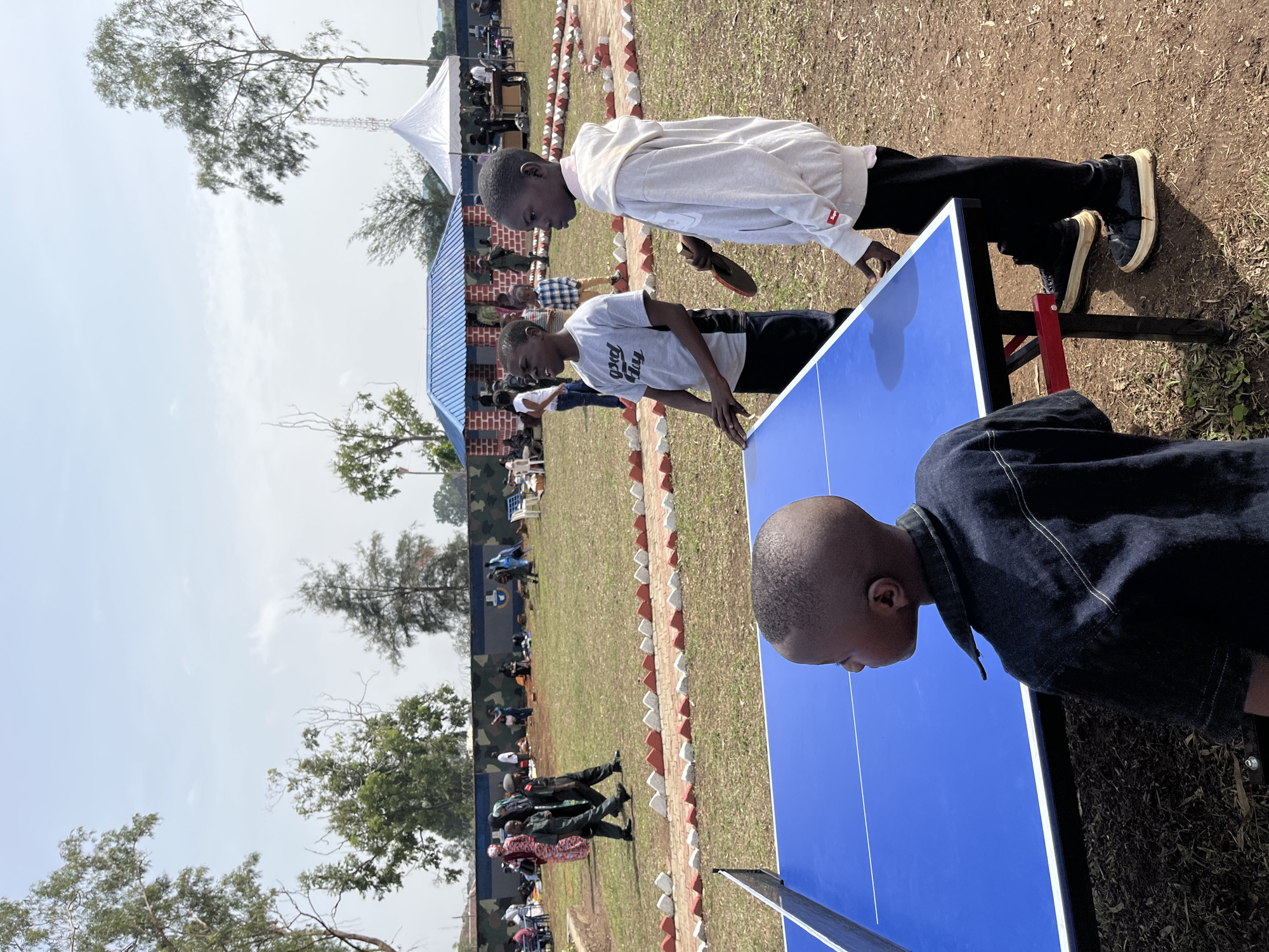 In Barakin Lerawi, Nigeria, an outdoor event is underway. A bright blue ping pong table is positioned on a dirt and grass surface. Two young boys are engaged in a game of ping pong, with one boy, wearing a grey t-shirt and black pants, actively playing, and the other, in a white hoodie and black pants, holding a ping pong paddle. Another young boy, seen from behind and wearing a dark denim shirt, stands to the left of the table. In the background, a few other individuals are visible, including two men in military-style uniforms walking together. Further back, a blue-roofed building with red brick accents stands under a clear sky. Tents are also present in the background. The event appears to be taking place in an open, grassy field with trees scattered around the perimeter.