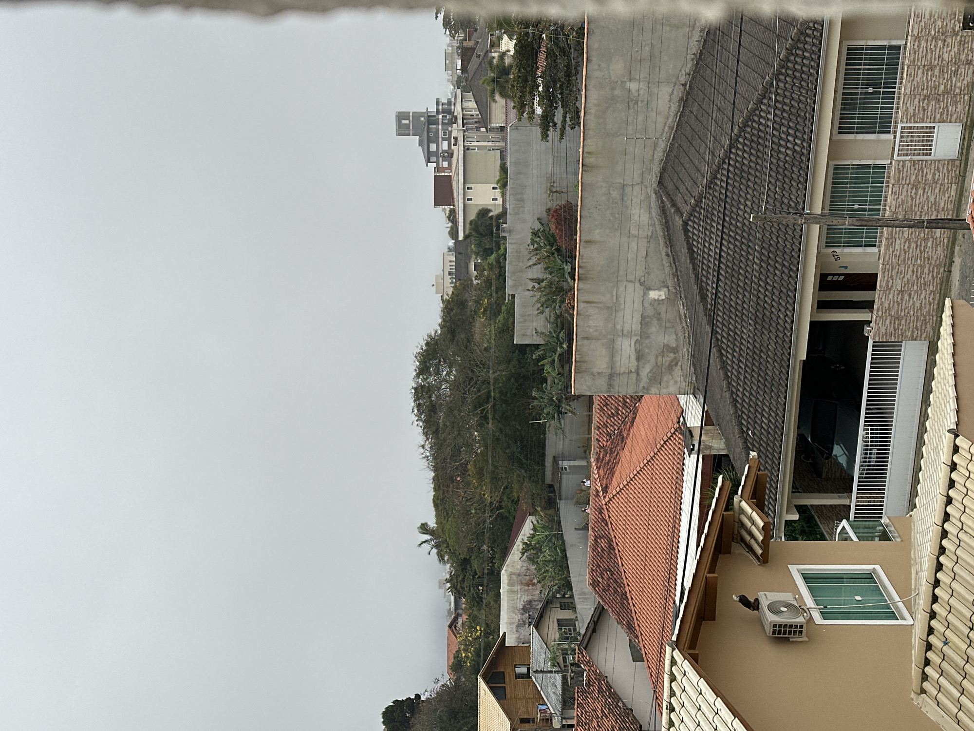 This image captures an overcast day over a residential area in Florianópolis, Brazil. The scene is dominated by rooftops of various houses and buildings, interspersed with dense green foliage. In the foreground, a light brown house with a white-tiled roof has an air conditioning unit mounted on its exterior wall. A dark-colored bird, possibly a hawk or eagle, is perched on the edge of a smaller roof above the air conditioning unit.
Moving towards the middle ground, several houses with orange-tiled roofs are visible. A concrete wall runs horizontally, separating different levels of the settlement. Beyond this, a modern multi-story building with a distinctive square tower on top stands out against the hazy sky. Further in the background, the landscape appears hilly and covered in trees, with more buildings nestled among them. The overall atmosphere is subdued due to the cloudy weather, and the image conveys a sense of a quiet, established neighborhood.