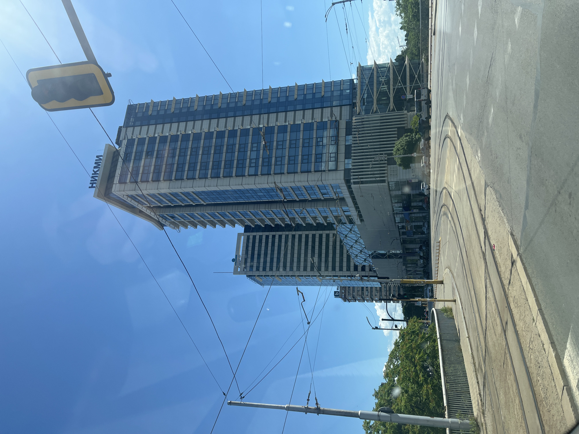 A wide-angle shot captures a modern cityscape under a clear blue sky. In the foreground, tram tracks curve across a paved road, indicating active public transportation. A low, metal-railed bridge or walkway borders the left side of the road, with lush green trees visible beyond it.
Dominating the background are several tall, contemporary buildings. The most prominent is a sleek skyscraper with a glass facade, topped with a sign reading "НИКМИ". Adjacent to it is another tall building with a similar architectural style. To the left and further in the background, other buildings of varying heights and designs are visible.
Numerous overhead wires, likely for the tram system, crisscross the sky, creating a network of lines against the blue. A traffic light with a yellow casing hangs suspended from a metal arm, also supported by wires. The overall scene suggests a busy urban environment with a blend of modern architecture and essential infrastructure. The lighting indicates it is daytime.