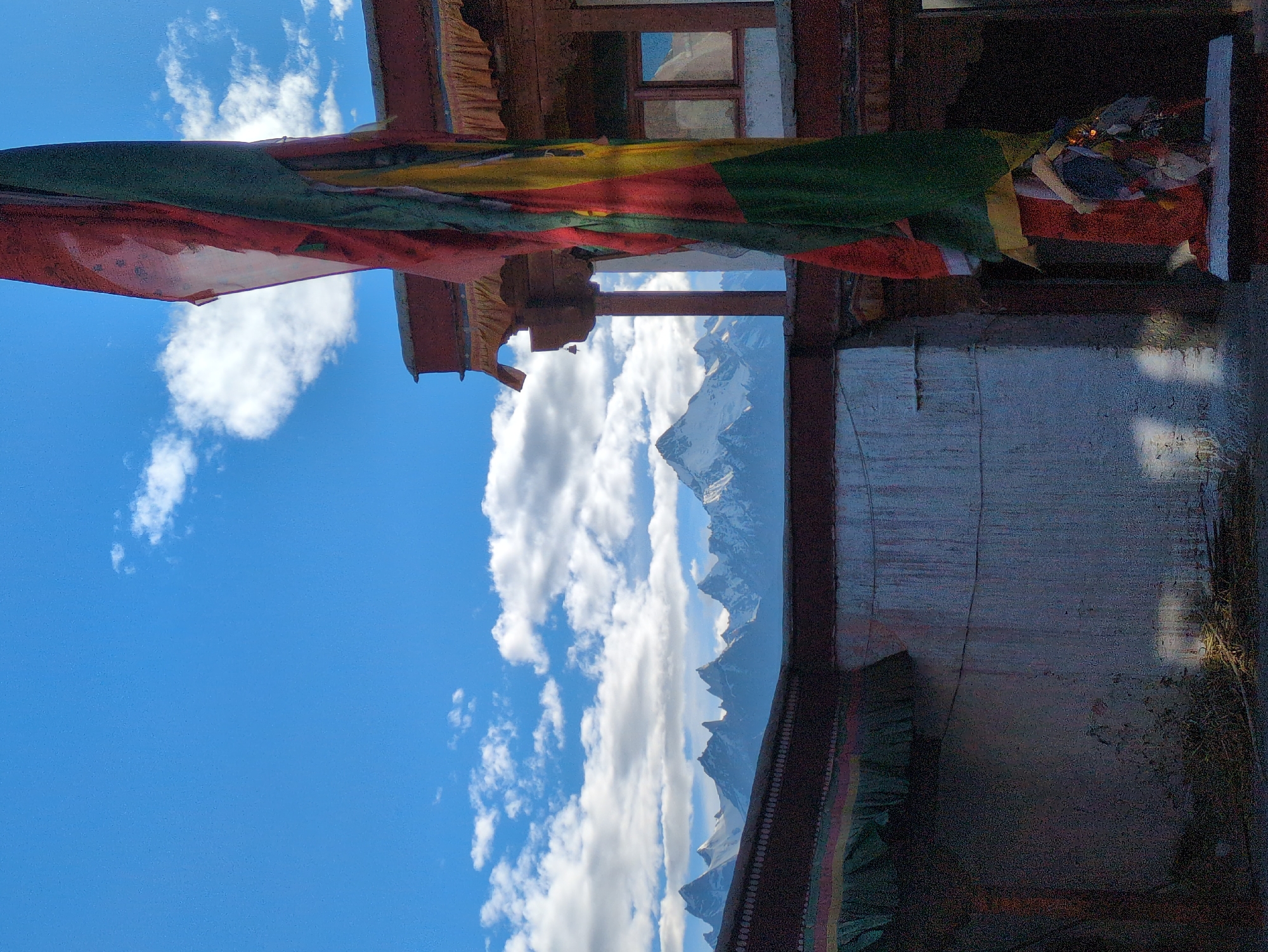 A prayer flag flutters in the foreground, with a building and snow-capped mountains in the background under a blue sky with clouds. The building appears to be a monastery or temple, with traditional architecture featuring wooden beams and decorative trim. The mountains are rugged and imposing, with snow covering their peaks. The prayer flag is made of multiple colored panels, likely representing the five elements of Buddhism. The overall scene suggests a peaceful and spiritual setting in a mountainous region. The location is identified as Yulang, India, a place known for its Buddhist monasteries and stunning natural beauty.