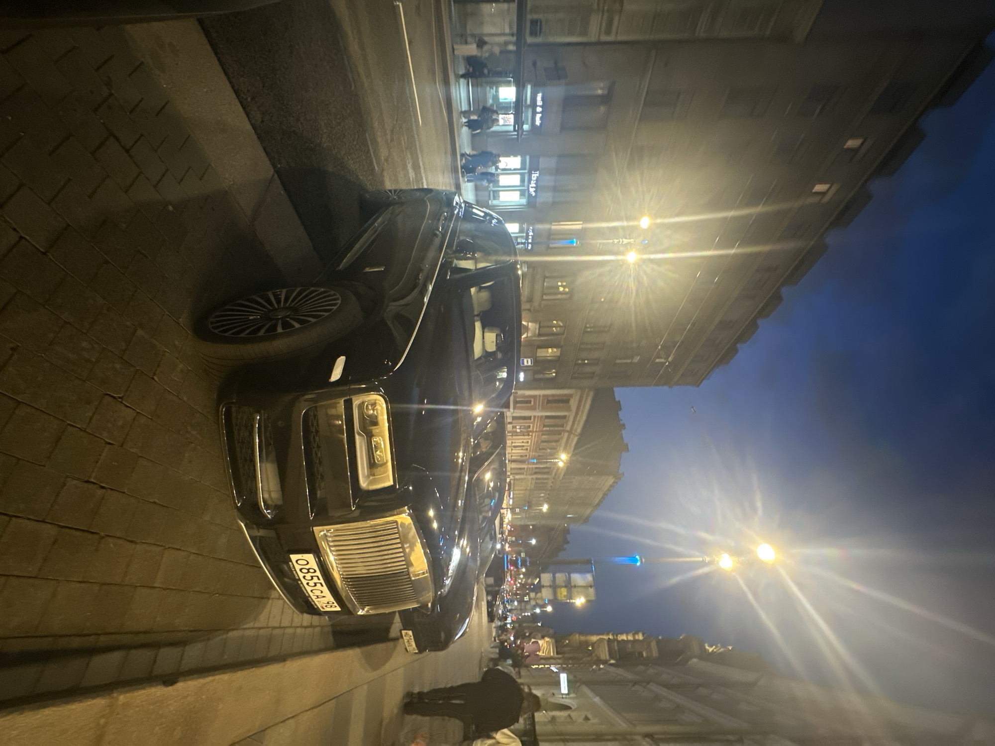 A black Rolls Royce is parked on a cobblestone street at night in Saint Petersburg, Russia. The car's front passenger door is open. The license plate number is O855CA 98. Pedestrians are walking on the sidewalk. The street is lined with buildings and streetlights. The sky is dark blue.