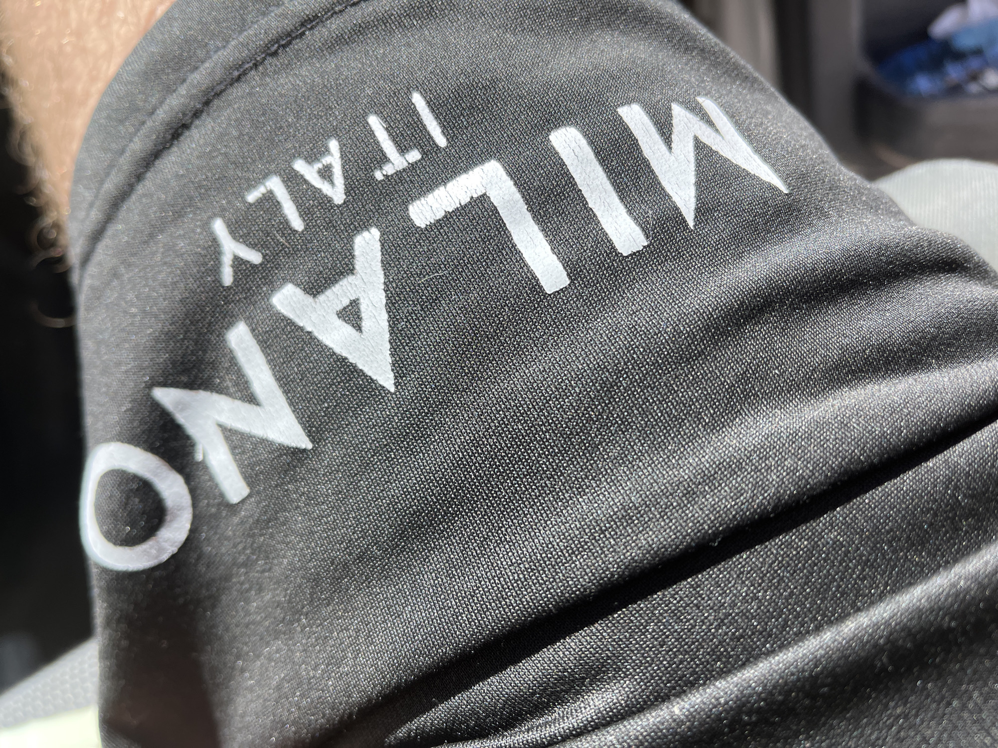 The image shows a close-up of a black garment with white lettering that reads "MILANO ITALY" vertically. The fabric has a textured, woven appearance. The letters appear slightly distorted due to the fabric's contours.