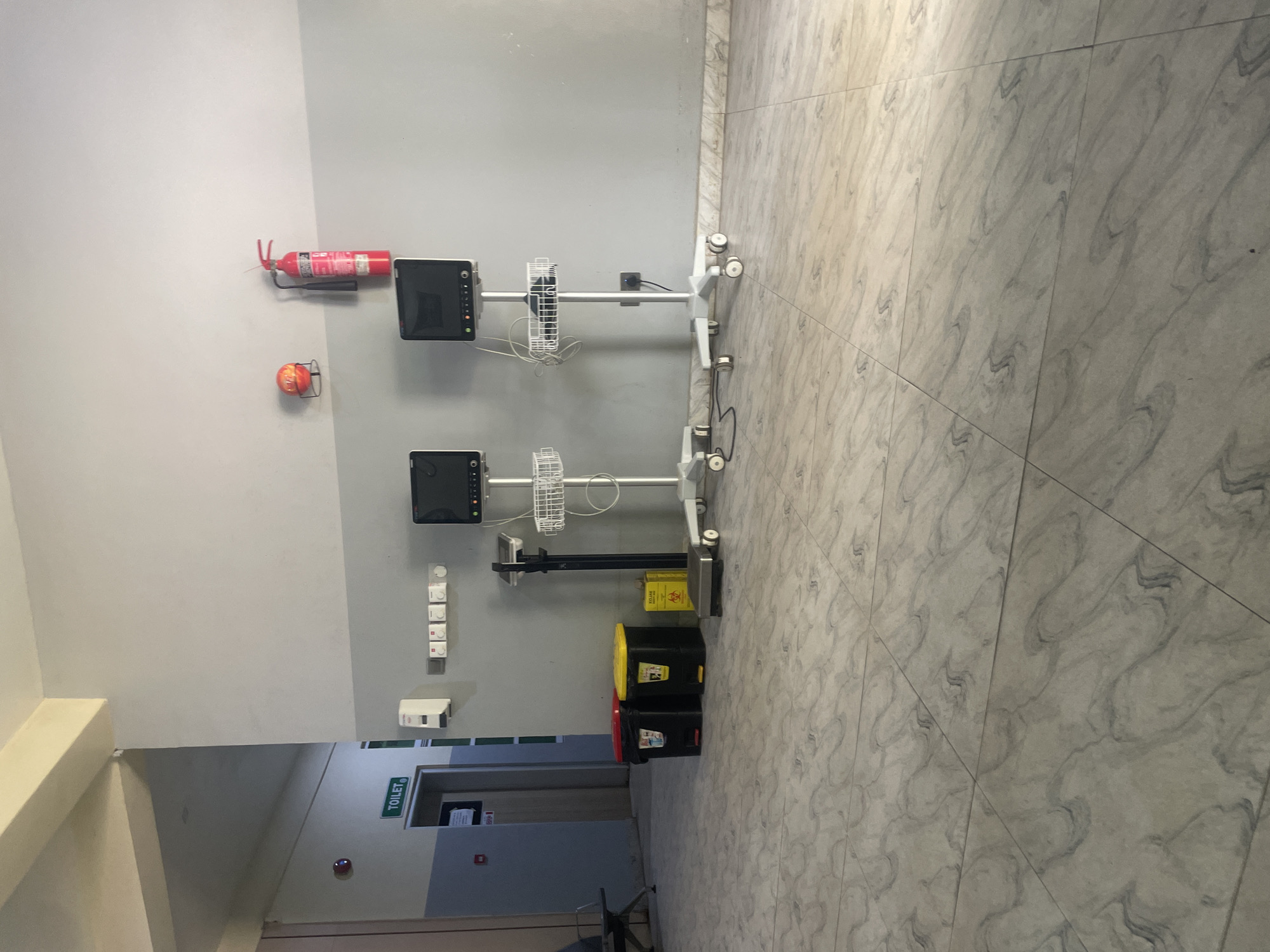 The image depicts a hallway within a medical facility, likely in Abuja, Nigeria. The hallway features a tiled floor with a marbled pattern. On the left, a sign with the word "TOILET" is visible above a doorway. Further down the hallway, a red emergency light is mounted on the wall.
The main focus of the image is the right side of the hallway. Here, two medical monitoring machines on rolling stands are positioned against a grey and white segmented wall. Above these machines, a red fire extinguisher and a circular orange object are mounted on the wall. To the left of the monitoring machines, a medical scale stands next to two waste bins, one red and one yellow, and a yellow biohazard disposal container.
Several electrical outlets and light switches are visible on the wall. The environment appears to be clean and functional, consistent with a healthcare setting. No people are present in the image. The lighting suggests it is daytime, with natural light possibly entering from an unseen source.