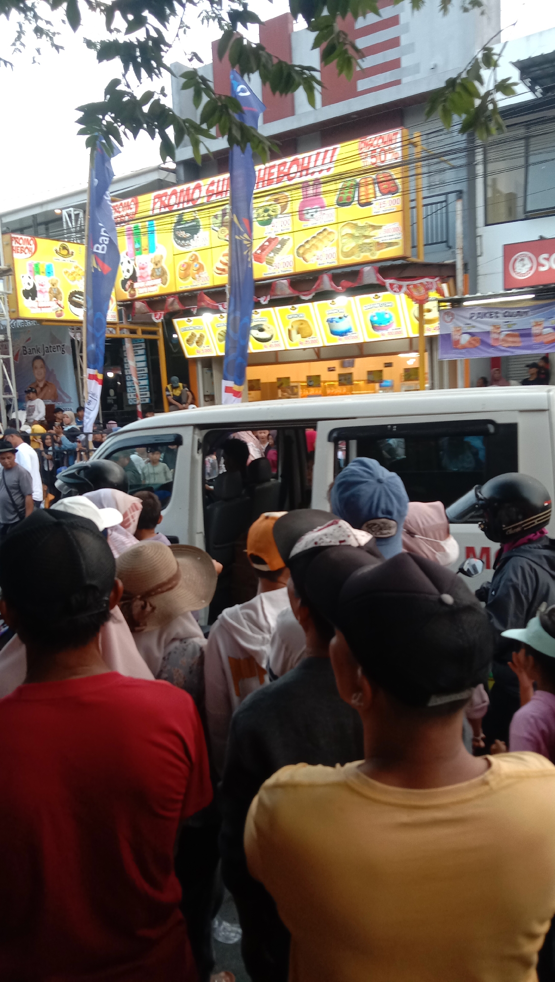 The image depicts a street scene in Jatisawit, Indonesia, with a crowd of people gathered in front of food stalls. A white van is parked in the foreground, with people entering and exiting. The stalls are decorated with colorful banners and display various food items, including cakes, pastries, and drinks. The overall atmosphere appears to be festive and lively, with a sense of community gathering. The presence of food stalls and the van suggests a possible event or market day.