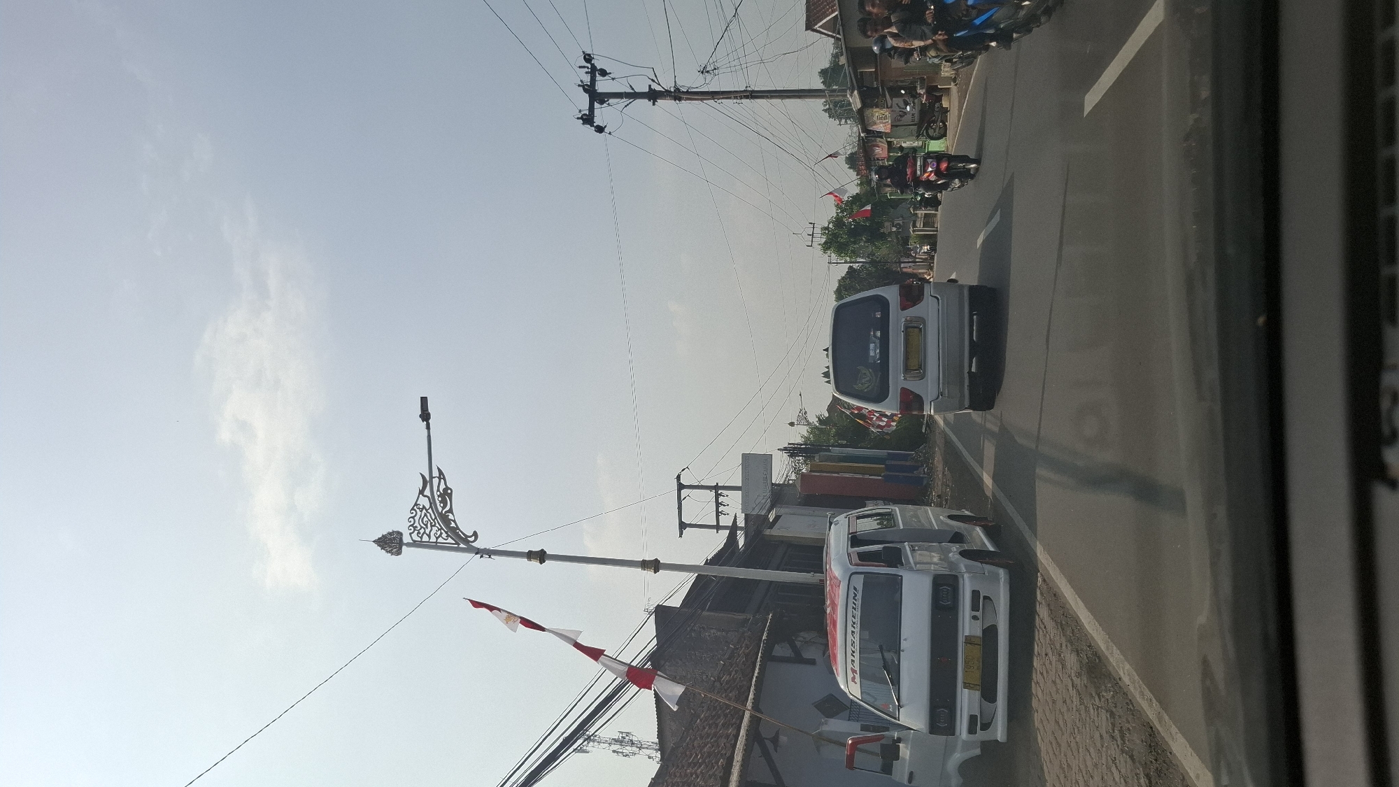 The image depicts a street scene in Cilaku, Indonesia. In the foreground, the dashboard of a vehicle is visible, indicating the photo was taken from inside a car. The road stretches ahead, with traffic including several white vans and motorcycles. On the left side of the road, a white van with red and white lettering is parked. Further down the street, a white van is driving away from the camera. On the right side of the road, several motorcycles are visible, some with riders.
The environment features a clear sky with scattered clouds. Electrical poles and wires are prominent throughout the scene. Decorative streetlights with ornate metalwork are also present, some adorned with Indonesian flags. Buildings line the sides of the road, appearing to be shops or residences. The overall impression is a typical day on a suburban or semi-urban road in Indonesia, possibly during a celebratory period given the flags.