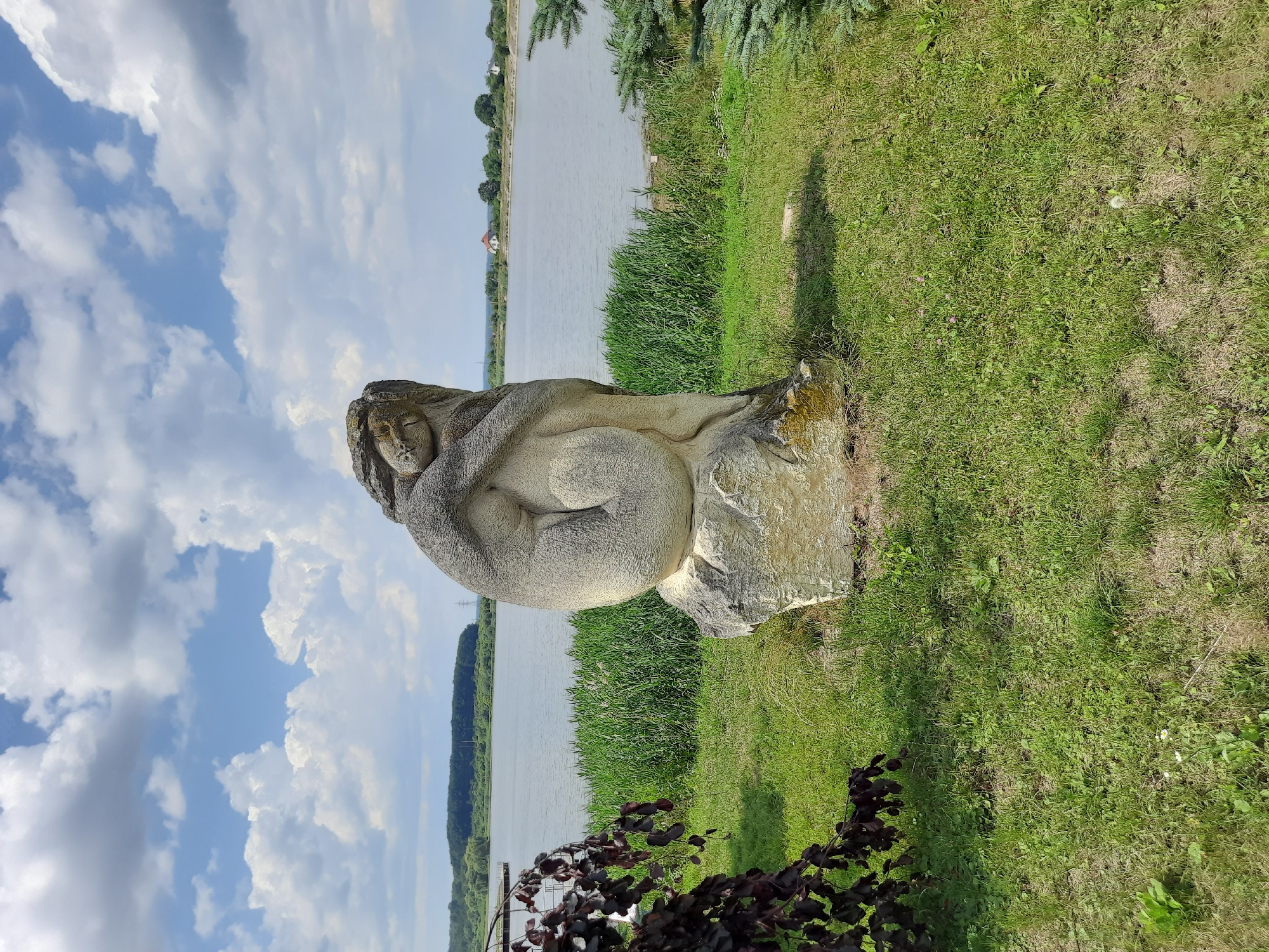 The image depicts a stone sculpture of a woman curled up, resting on a large boulder. The sculpture is situated outdoors, on a grassy area next to a body of water, possibly a lake or reservoir. In the background, there is a line of trees, and some houses and structures are visible on the opposite shore. The sky is partly cloudy with white clouds against a blue sky. A dark-leafed plant is in the foreground on the left side of the image, and a pine tree branch is visible on the right. This sculpture is located in Drohobych, Ukraine, specifically at the "Kamianna Zvukiv" (Stone Sounds) park, known for its stone sculptures.