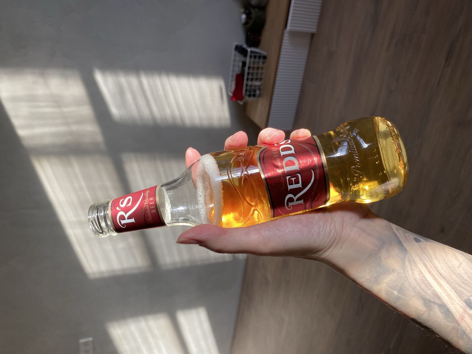 A hand is holding a partially consumed bottle of Redd's Premium Quality Beer. The bottle is tilted, with the neck pointing towards the left of the frame. The hand and the bottle are illuminated by sunlight. The background consists of a wall with sunlight patterns and a wooden floor. In the blurred background are a white basket, and furniture that could be drawers or shelves. There is no indication of this image being taken in the city of Votkinsk, Russia.