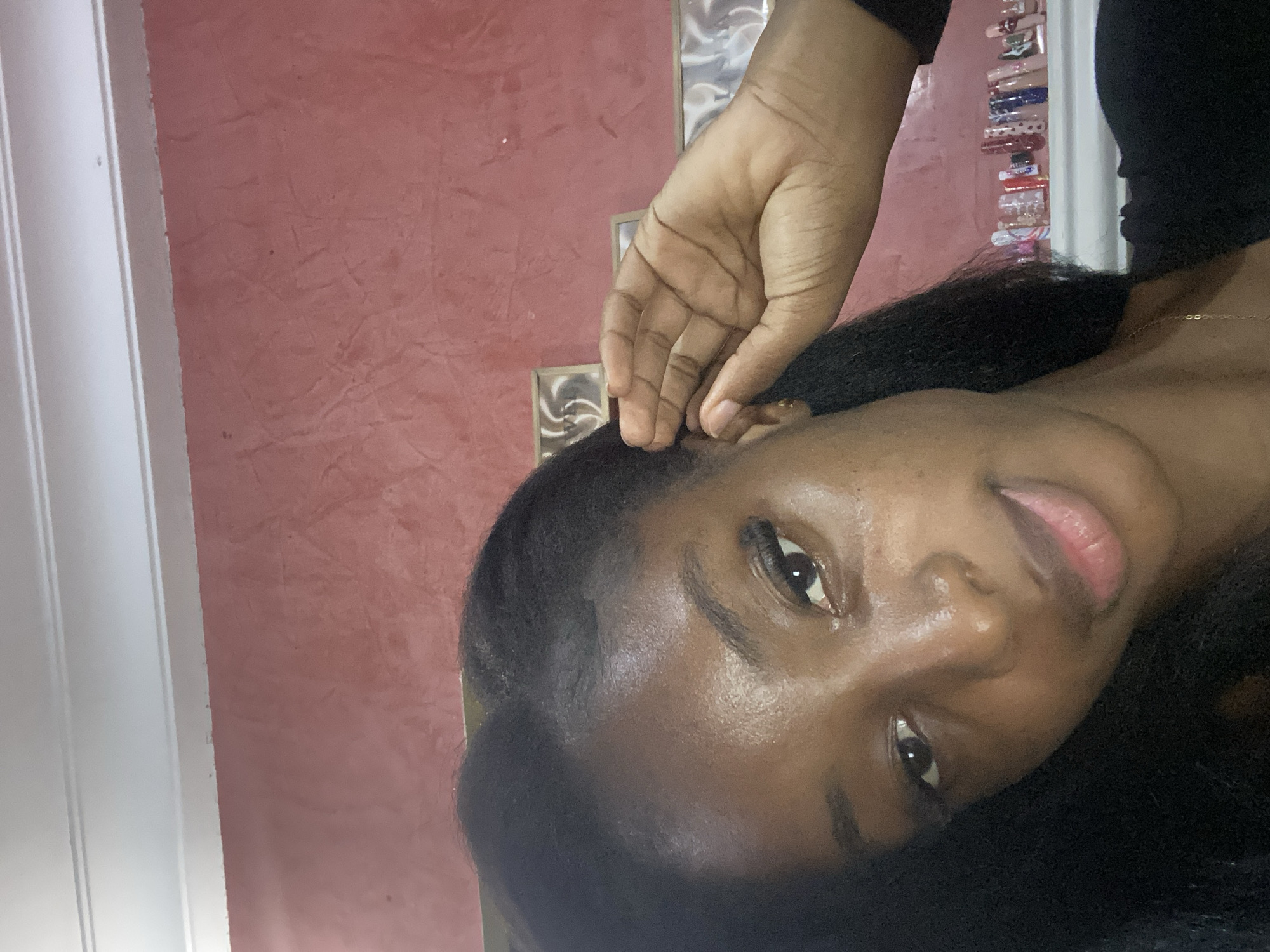 The image shows a close-up of a young Black woman's face, taken from a slightly low angle. She has dark skin, dark eyes, and long, dark hair that is partially obscuring her face. Her left hand is raised and gently touching her ear. She is wearing a black top and a delicate gold necklace. The background is a textured red wall with white molding at the top. On a shelf behind her, there are various items, including what appear to be nail polish bottles and small framed pictures. The lighting suggests an indoor setting. The image does not contain any specific visual cues that would indicate its location in Jos, Nigeria.