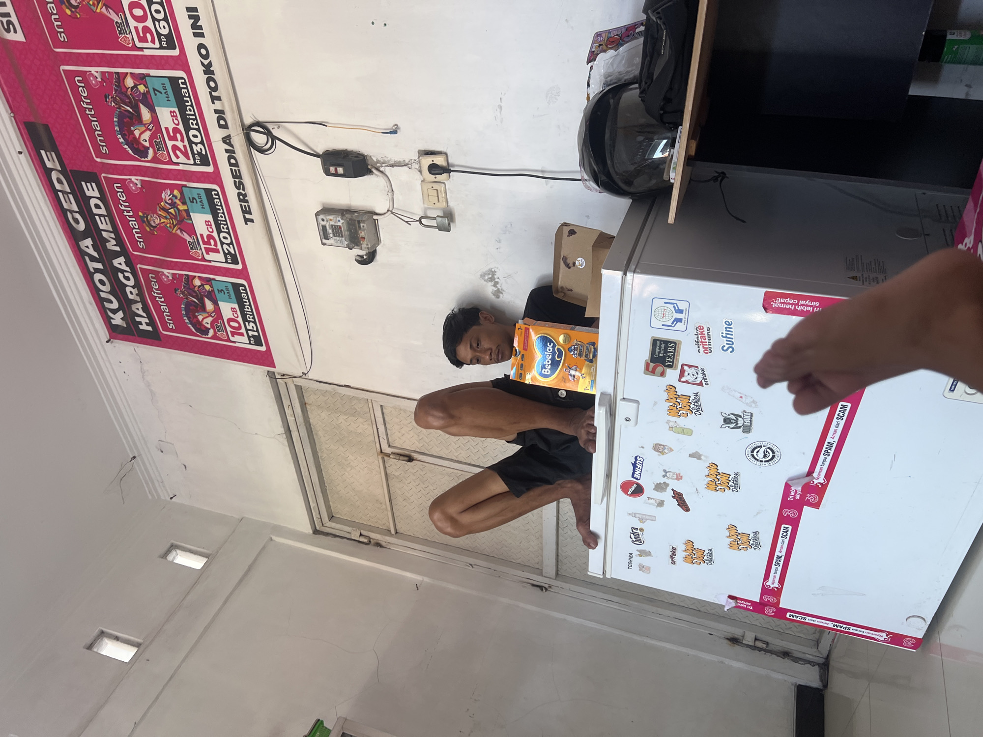 In a room in Kecamatan Pati, Indonesia, a young man is sitting on top of a white refrigerator. He is wearing black shorts and a black t-shirt, and his legs are bent at the knees, with his feet pointing upwards. He is holding a box of baby formula, "Bebelac," and looking downwards. The refrigerator is covered in various stickers, including brands like "Toshiba," "Candra," and "Orifake," as well as promotional tapes for "Smartfren." Above the refrigerator, a pink banner advertises "KUOTA GEDE HARGA MEDE" (Big Data, Moderate Price) with offers for Smartfren mobile data packages. Electrical outlets and a padlock are mounted on the wall behind the man. To the right of the refrigerator, there is a shelf with a black motorcycle helmet and other items. A hand and foot are visible in the foreground, suggesting the perspective of someone taking the photo. The room appears to be a small shop or a domestic space.