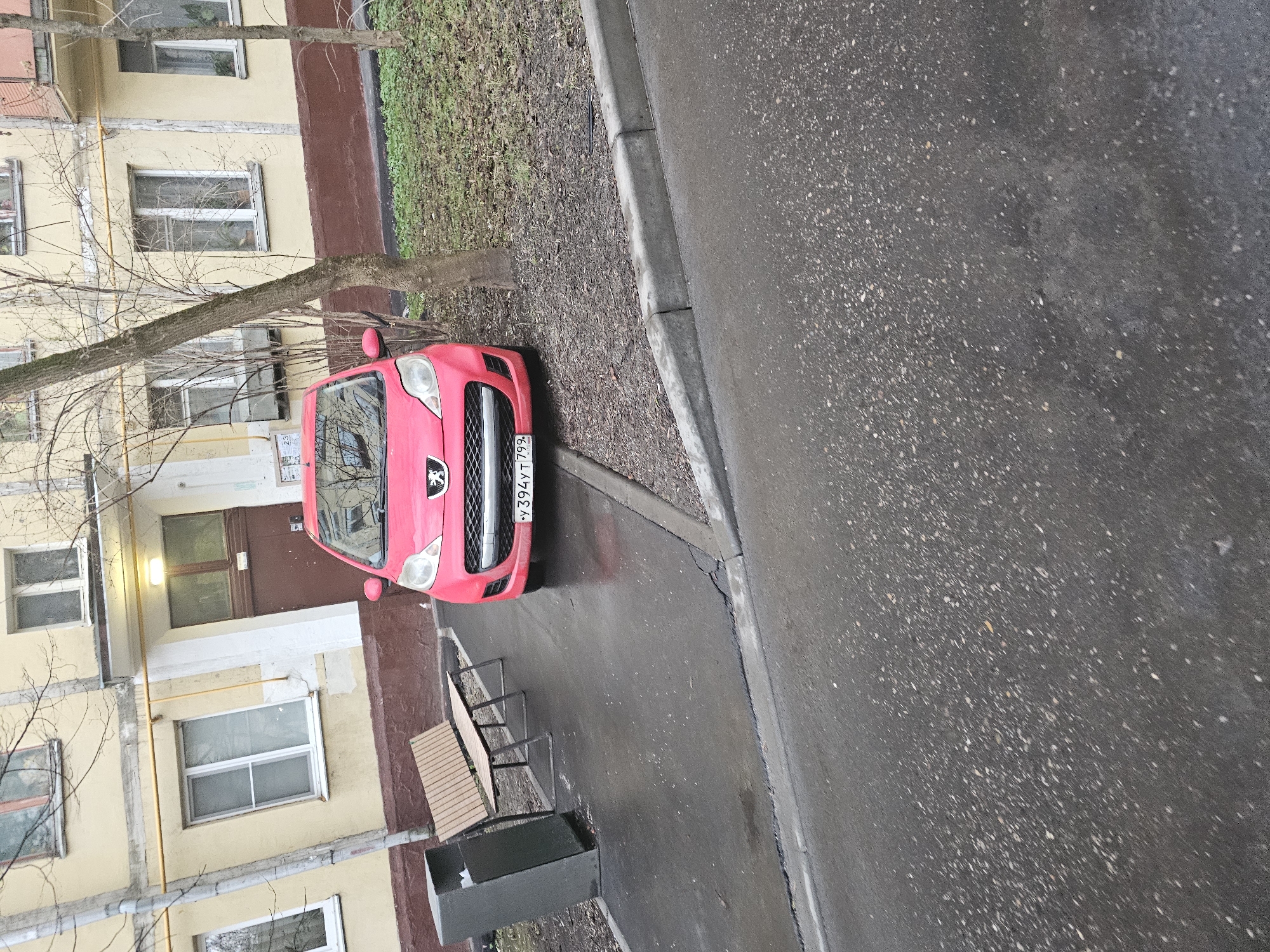 The image depicts a red Peugeot car parked adjacent to a residential building on a wet paved surface. The building appears to be multi-storied, with beige facade and windows. There's a small patch of vegetation between the building and the parking spot. A bench and a trash receptacle are visible beside the parked vehicle. The car's license plate is partially visible, reading "Y394YT 799". The location is possibly in Khimki, Russia, given the Russian license plate and building style. No people are visible in the image.