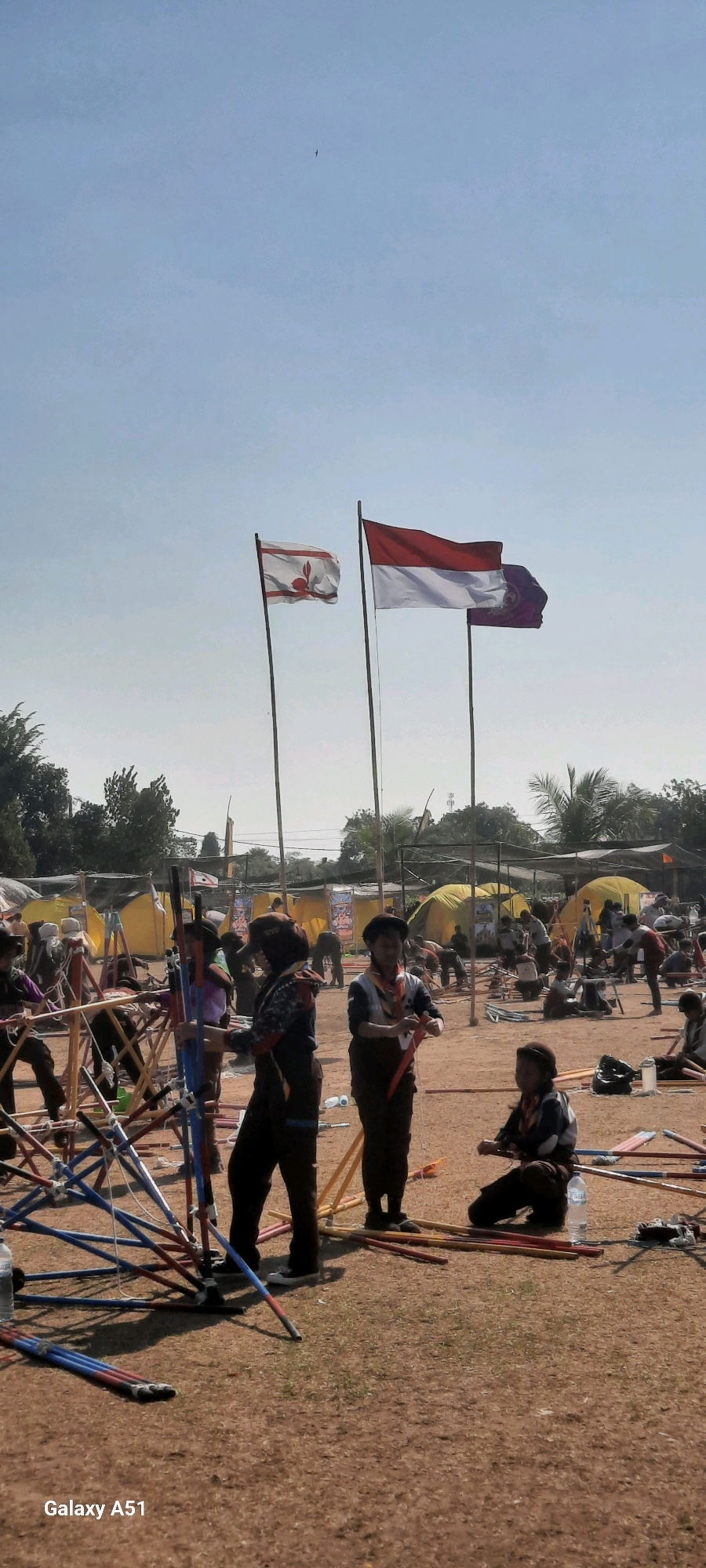 The image depicts an outdoor event, likely a scout jamboree or similar activity, taking place on a dirt field in Selopuro, Indonesia. The sky is clear and pale blue, with a single bird visible in the distance. Two flags are prominently displayed on tall poles in the mid-ground; one is red and white, resembling the Indonesian flag, and the other is white with a red emblem. In the foreground and mid-ground, numerous individuals, predominantly young people in scout-like uniforms, are engaged in various activities. Some are assembling or disassembling structures made of colorful poles and ropes, possibly for tents or camp setups. Others are standing or sitting, observing or holding materials. Yellow tents are visible in the background, suggesting a temporary encampment. The ground is covered in dry earth and sparse patches of grass, with scattered poles and equipment lying around. The overall atmosphere appears to be one of organized activity and outdoor recreation.