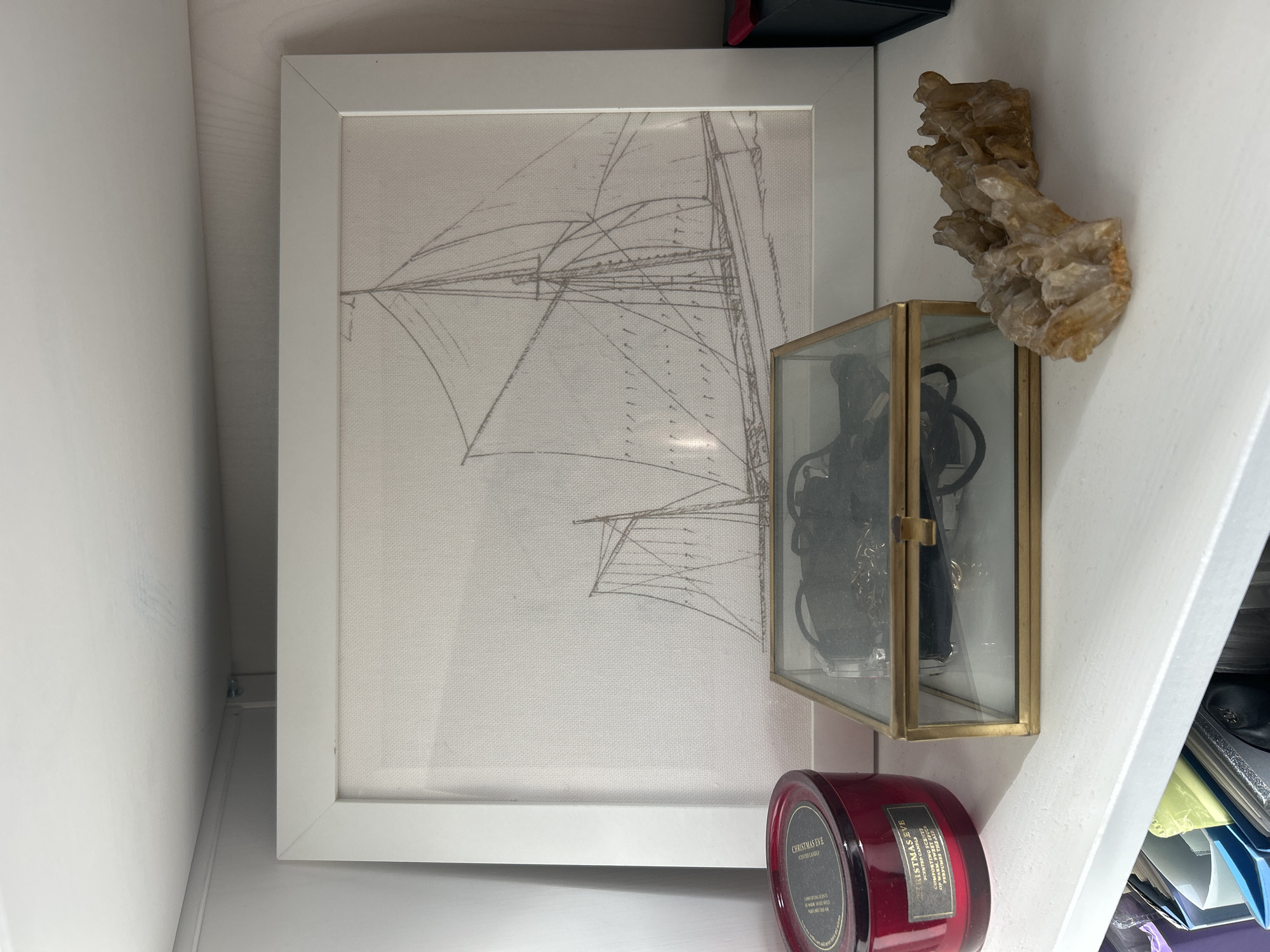 This is a close-up shot of a shelf in what appears to be a closet or storage area. The main subject is a framed sketch of a sailing ship. The sketch is rendered in thin, gray lines on a light-colored fabric background, and it depicts the ship's rigging and sails. The frame is a simple white border.
To the right of the framed sketch, there's a small, clear glass and gold-trimmed box. Inside the box, several dark-colored items, possibly jewelry or accessories, are visible. Above the glass box, a cluster of light-brown, crystalline formations is attached to the white wall, resembling a piece of raw quartz or similar mineral.
Below the glass box, a deep red scented candle is placed on the shelf. The candle is in a glass jar with a label that reads "CHRISTMAS EVE SCENTED CANDLE" and lists "COMFORTING SCENTS OF MULLED WINE, FIR NEEDLES AND CINNAMON."
The overall setting is indoors, with white painted walls and shelves. The lighting appears to be artificial, casting soft shadows. There are no people or animals visible. The image has a clean, minimalist aesthetic, with a focus on the decorative objects displayed on the shelf. The time of day and weather are not discernible from the image, nor are specific location cues beyond it being an indoor storage space.