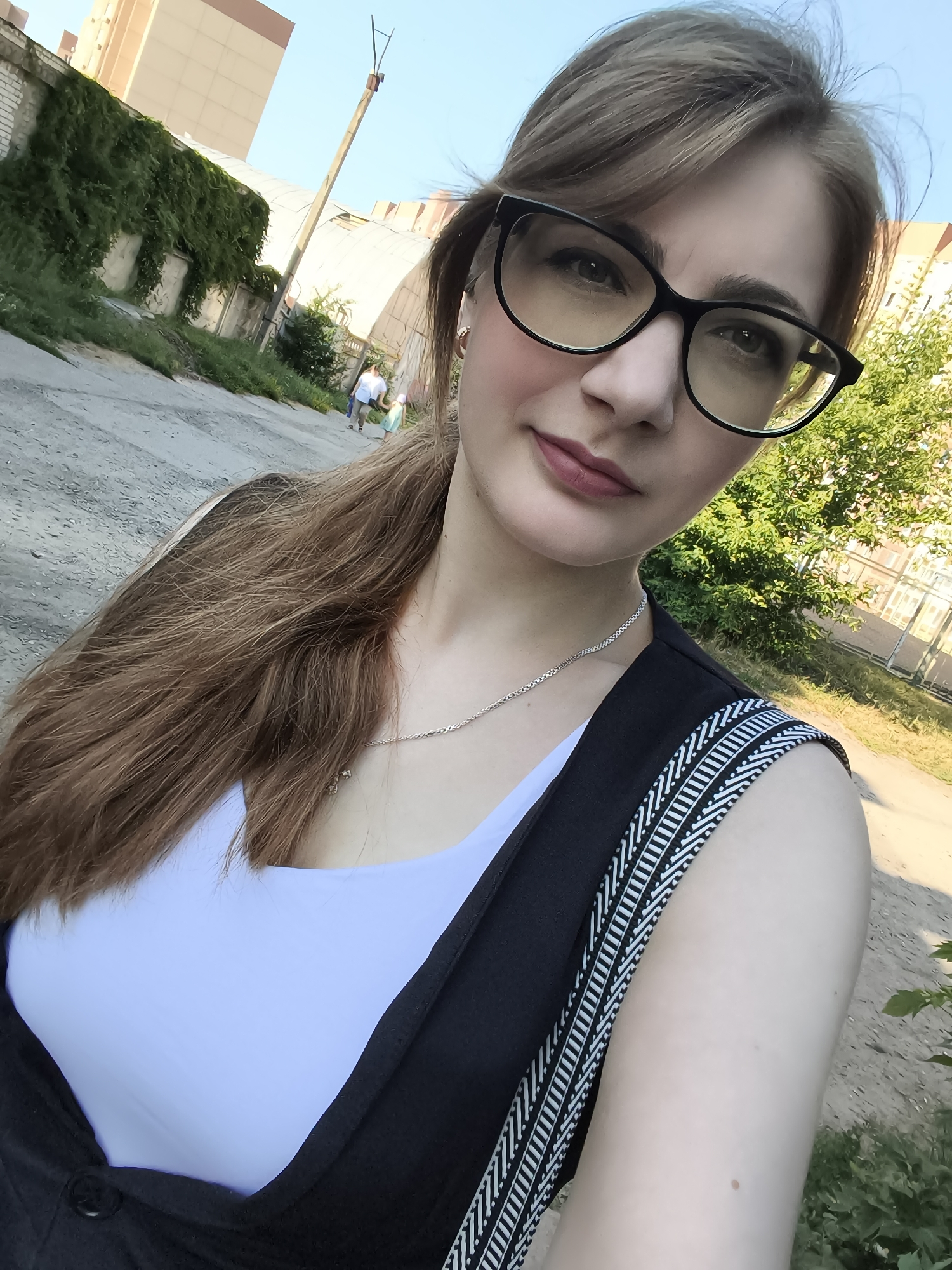 A young woman with long brown hair, wearing glasses, a white top, and a black vest, takes a selfie outdoors. She has on a silver necklace. In the background, a paved path winds past buildings covered in ivy and trees. Two people, possibly a parent and child, are walking away from the camera in the distance. The sky is blue and clear, suggesting it is daytime with fair weather. The scene appears to be in a residential area in Podkletnoe, Russia. The woman's expression is neutral, and the overall mood is casual.