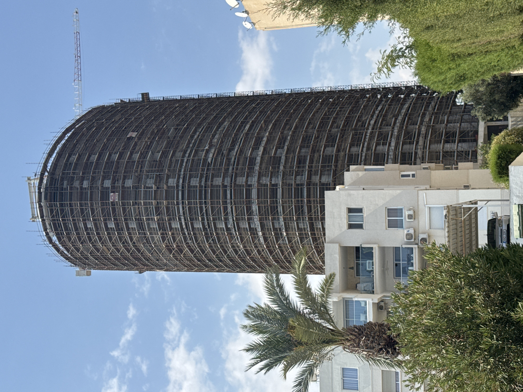 This image shows a cylindrical high-rise building under construction in the Agios Tychona Community, Cyprus. The building is extensively covered in scaffolding. A crane arm is visible extending from the top of the building towards the right side of the frame, with its base obscured by the scaffolding. In the foreground, to the left, a palm tree stands next to a white multi-story residential building. This residential building has balconies with glass railings and windows, and air conditioning units are visible on its facade. A dense green bush is in front of this building. To the right of the high-rise, another building with satellite dishes on its roof is partially visible. The sky is blue with scattered white clouds. The scene is illuminated by daylight. No people are discernible in the image.