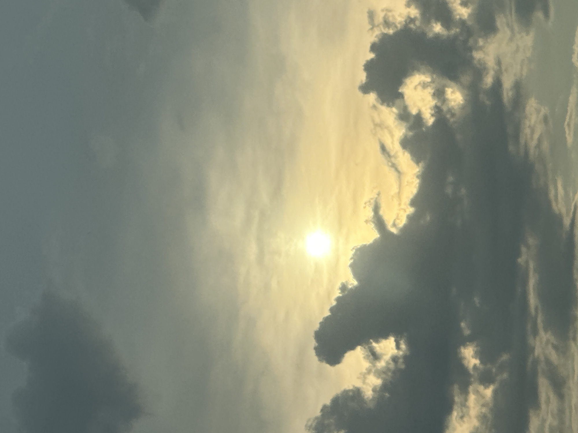 The image shows a sky with a bright sun peeking through dark, dramatic clouds. The sun is located in the upper center of the frame, casting a warm yellow and orange glow. The clouds are a deep grey, with some illuminated edges where the sunlight catches them. The overall impression is one of a dynamic and somewhat moody sky. There are no visible people, structures, or any recognizable landmarks that would indicate a specific location within the city of Skorotovo, Russia. The scene is purely atmospheric.