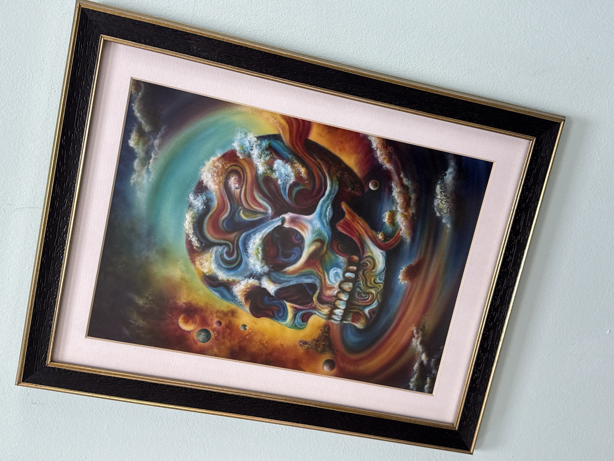 A framed painting hangs on a light blue wall. The painting depicts a skull with swirling colors of blue, red, and yellow, set against a dark background with planets and a rainbow. The painting has a black and gold frame with a light pink mat. The signature "S. Akan" is visible in the upper right corner of the painting. There is no evidence to suggest this image was taken in Burgas, Bulgaria.