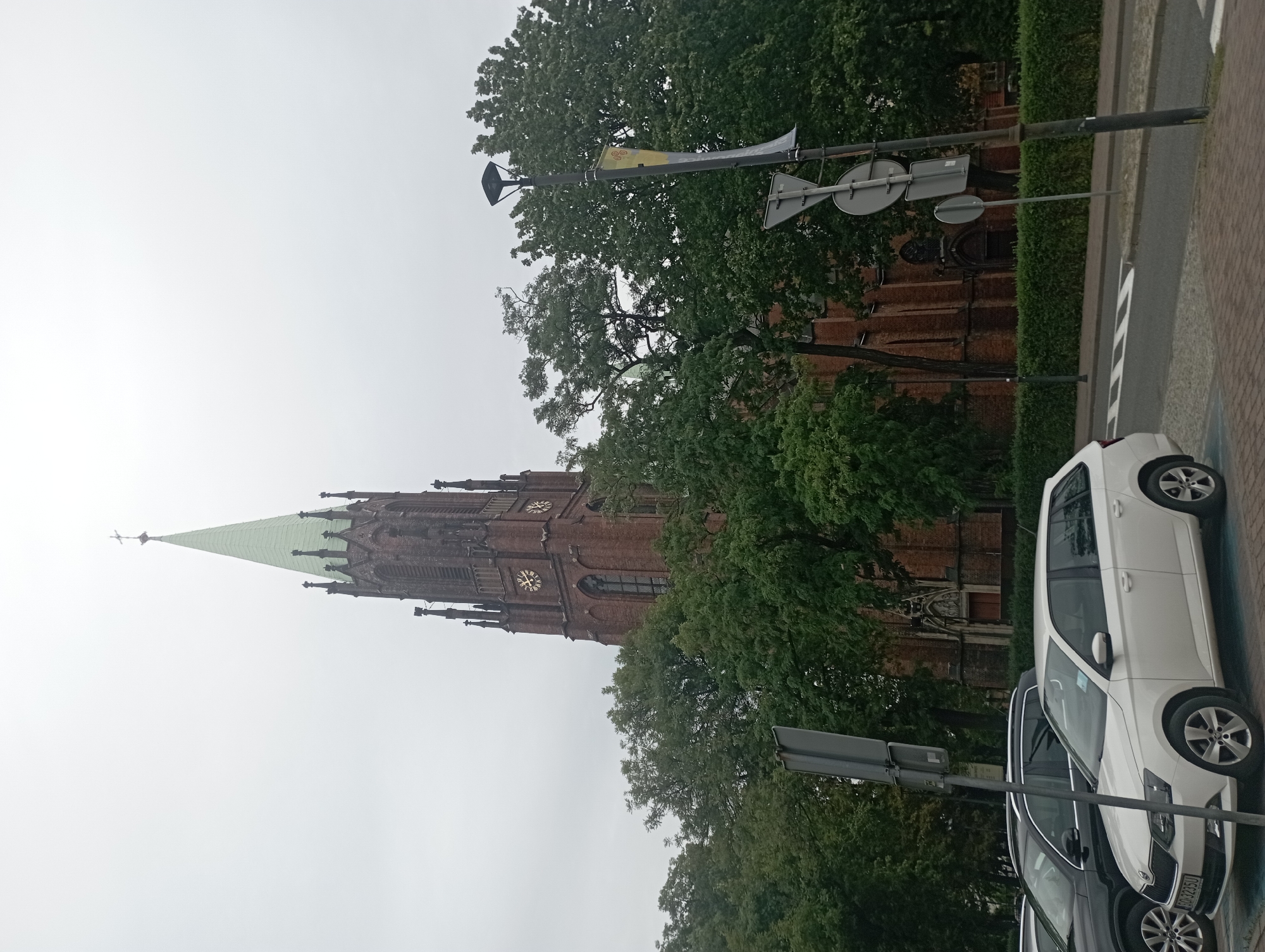 The image depicts the St. Mary's Assumption Church in Siemianowice Śląskie, Poland, under an overcast sky. The church's prominent feature is its tall, brick tower topped with a green spire, adorned with Gothic architectural elements and a clock face. The building's facade is made of red brick. Surrounding the church is a dense growth of lush green trees, partially obscuring the lower levels of the building. In the foreground, several cars are parked on a paved area adjacent to a road. A white station wagon is positioned closest to the viewer, with a dark-colored SUV behind it. Road signs and a lamppost are also visible near the vehicles, indicating a public urban setting. The overall scene conveys a sense of a typical cloudy day in a city, with a significant architectural landmark serving as the backdrop to everyday life.