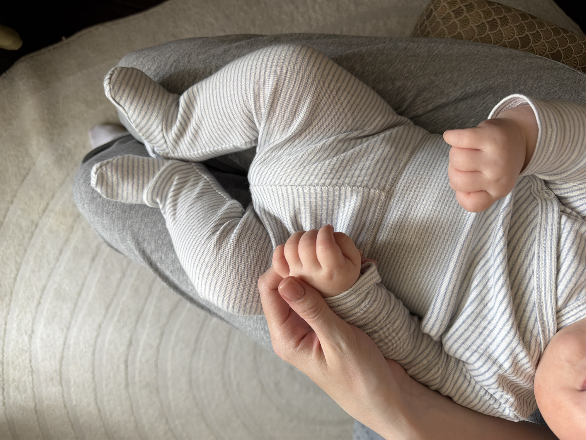 This overhead photograph captures a tender indoor scene in Khimki, Russia, featuring a baby resting on an adult's lap. The baby is dressed in light-colored footed pajamas with fine, light blue or grey stripes, covering their entire body including their feet. One of the baby's small hands is gently clasped by an adult's hand, while the other is curled into a fist. The baby's face is partially visible in the lower right corner of the frame.
The adult is wearing grey trousers or leggings, providing a soft surface for the baby. In the background, a light-colored rug or carpet with a subtle ribbed texture covers the floor. A patterned, darker object, possibly a cushion or blanket, is visible behind the adult's leg on the right side. The lighting appears soft and natural, suggesting it is daytime. The image evokes feelings of comfort, connection, and parental care. No text is visible in the media file.