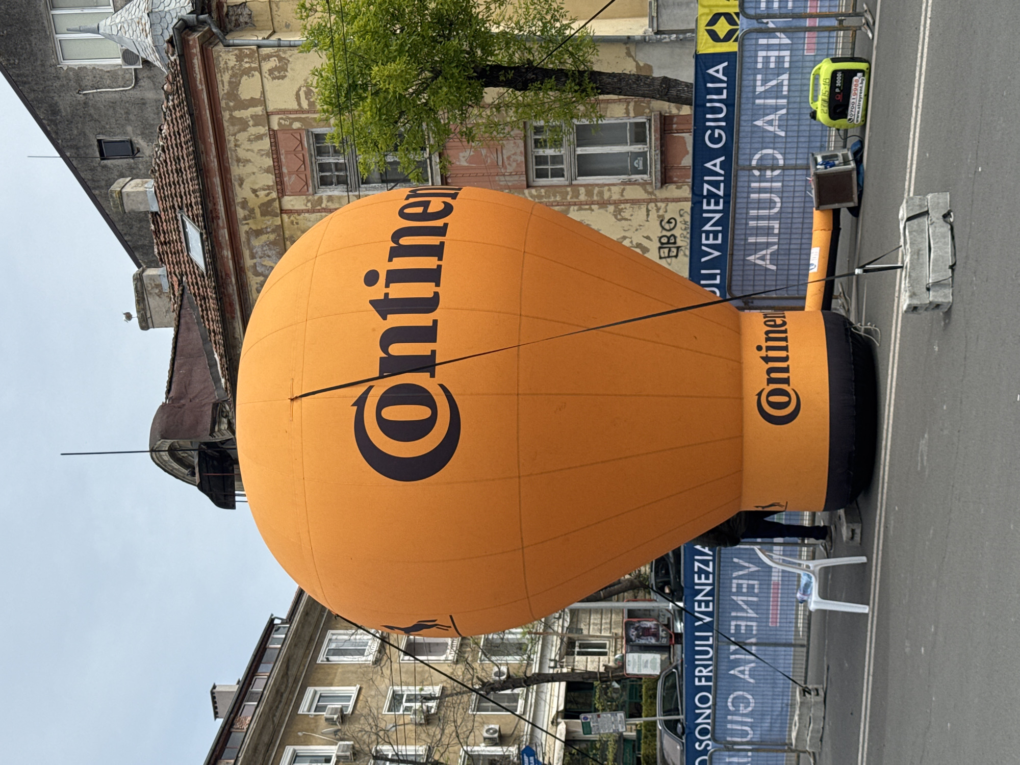 A large, orange, teardrop-shaped inflatable with the "Continental" logo dominates the scene, set against a backdrop of weathered buildings. The buildings feature windows, roof details, and peeling paint. A tree branch extends into the frame. Behind the inflatable, a blue banner reads "SONO FRIULI VENEZIA GIULIA" in mirrored text. A generator and equipment stand nearby. In the foreground, a white plastic chair sits on a paved surface. Based on the banner and architectural style, it is probable that the location is a European city. Without further information, it cannot be definitively confirmed that the location is Burgas, Bulgaria.