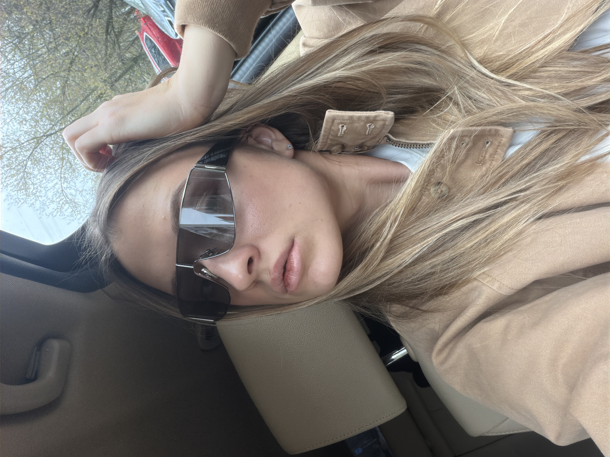The image, originally rotated, depicts a young woman in a relaxed pose, leaning her head against a car's headrest. She has long, straight, light brown hair with blonde highlights that frame her face and fall over her shoulders. She wears large, dark, reflective sunglasses that obscure her eyes, giving her a cool, calm demeanor. A small, sparkling stud earring is visible in her right ear.
She is dressed in a light tan or beige jacket, which appears to be made of a sturdy fabric like denim or canvas, worn over a white top. Her right hand is casually resting on her head.
The setting is inside a car, indicated by the light-colored, possibly leather, headrest and seat, along with a grab handle visible in the upper left. Through the car window, an out-of-focus background shows trees with new green leaves, suggesting springtime. A hint of a red vehicle is also visible in the reflection or distance. The scene is illuminated by natural daylight, indicating it is daytime. The mood is relaxed and casual, possibly a moment of rest or a posed photo while traveling. No visible text is present in the image. The location context provided is Moscow, Russia.