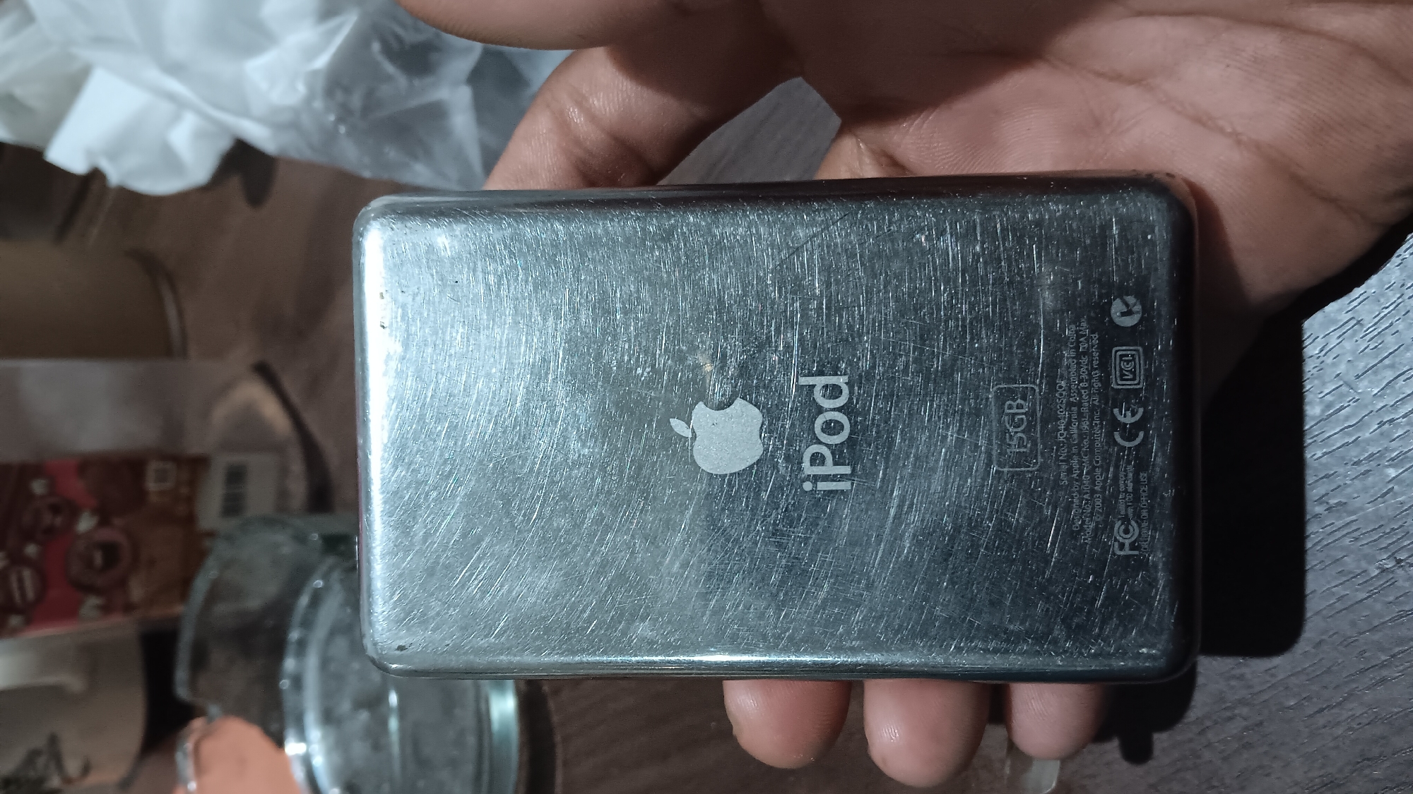 An extreme close-up, low-angle shot shows the back of a heavily scratched, silver colored iPod Nano. The iPod is being held by a person's fingers on the left and right sides of the frame. The Apple logo and the word "iPod" are visible in the center of the iPod. Below "iPod" is a small text indicating "15GB". Further down, more text is visible, including "Serial No: JQ401025QOF" and "2003 Apple Computer, Inc. All rights reserved.". The background is blurred, but suggests an indoor environment with a wooden table surface and other out-of-focus objects, including what appears to be a glass bowl on the left. The image does not provide any specific geographic information that would place it in Depok, Indonesia.