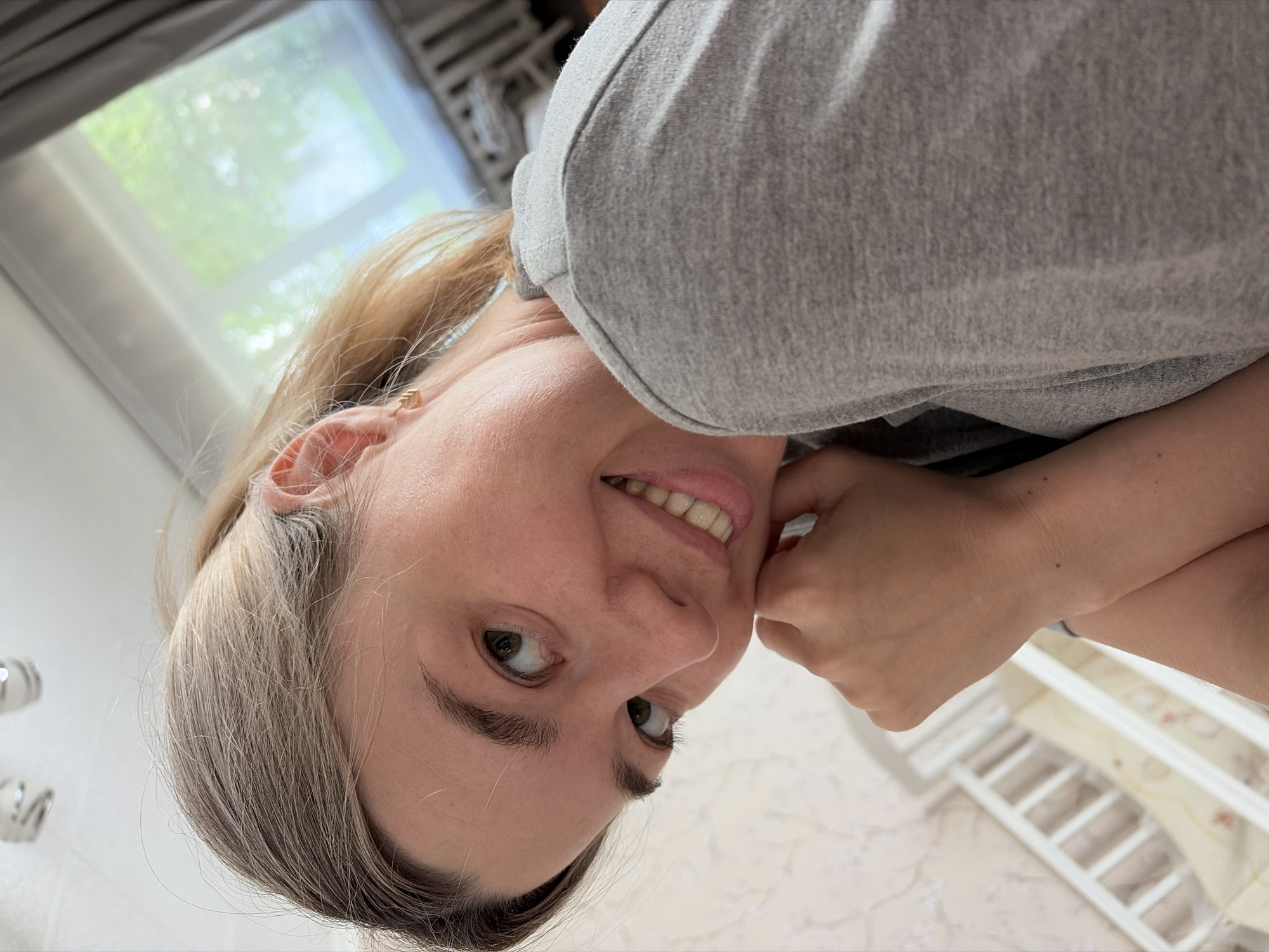 A selfie shot depicts a woman with light skin, graying blonde hair pulled back, and brown eyes. She is smiling slightly, resting her face on her hand. She wears a gray shirt. In the background is a window with green trees visible outside, and a white crib or railing. The location is the city of Chaikovsky, Russia.