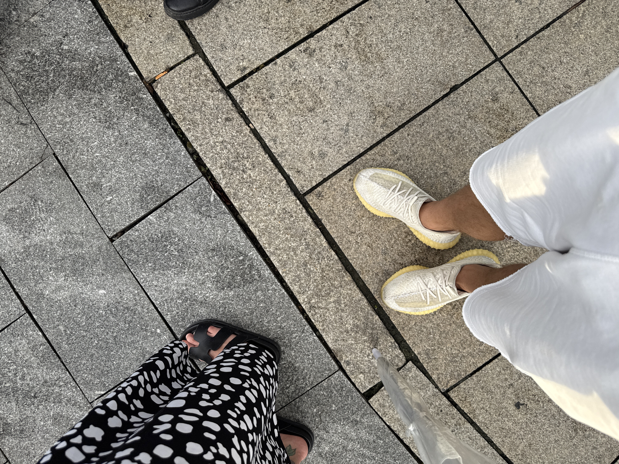 Two people are standing on a pedestrian walkway made of granite paving stones. One person is wearing black sandals and a black and white polka-dot skirt. The other person is wearing white sneakers and white pants. The perspective is from above, looking down at their feet and legs. In the lower right corner, a partially visible clear umbrella rests on the ground. The scene suggests an outdoor setting, possibly a city street or park, given the paving and the presence of people. The lighting indicates it could be daytime, perhaps late afternoon or early morning, judging by the soft shadows. The overall mood appears casual and everyday. There is no visible text in the image.
