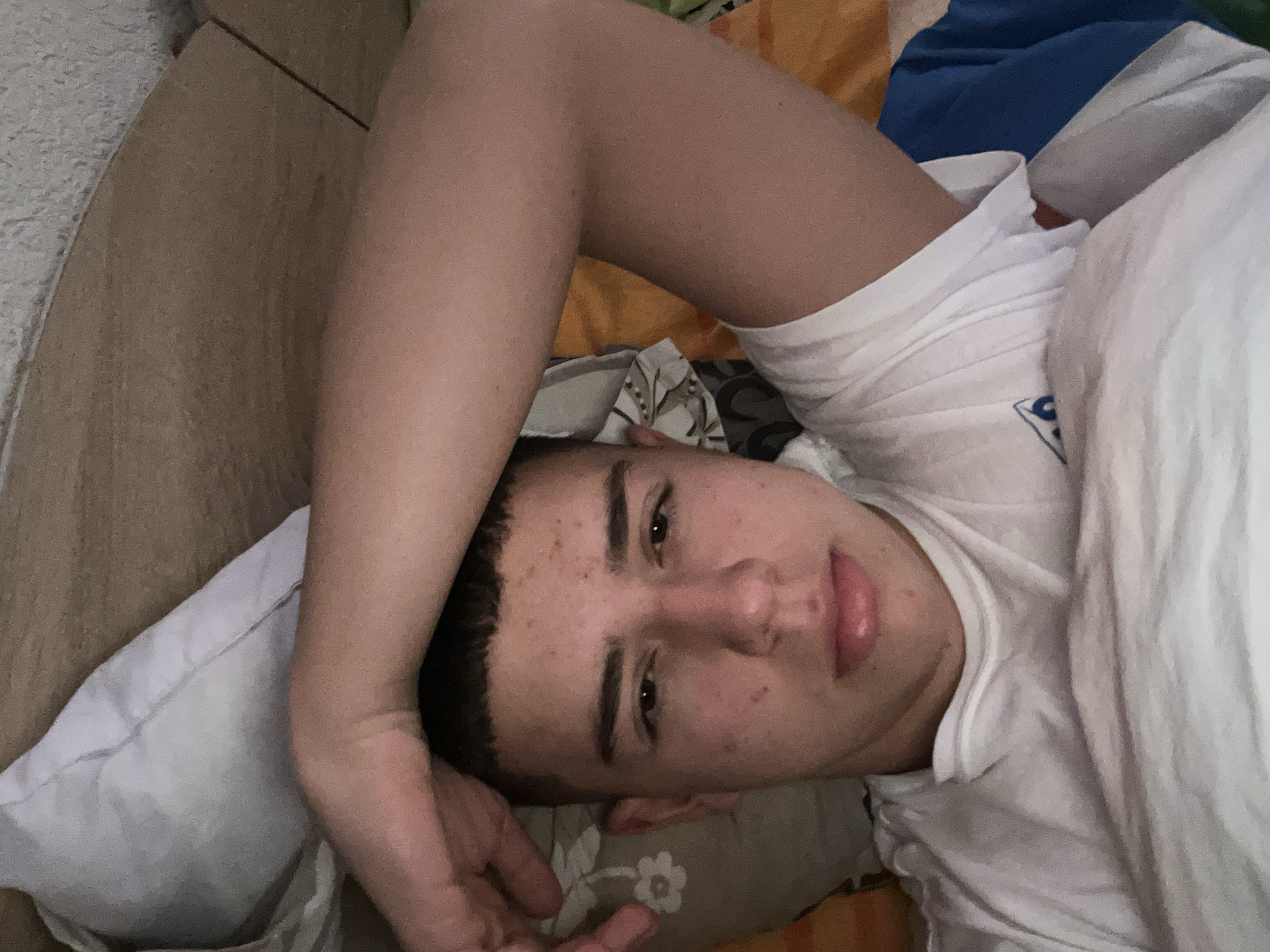 A young man is lying on a bed. He is fair-skinned, with short dark hair and dark eyes. He is wearing a white t-shirt. His head is resting on a white pillow, and his arm is bent over his head. The bed has a patterned sheet. The image appears to have been taken indoors. There is no verifiable information on the location in Zlatograd, Bulgaria.
